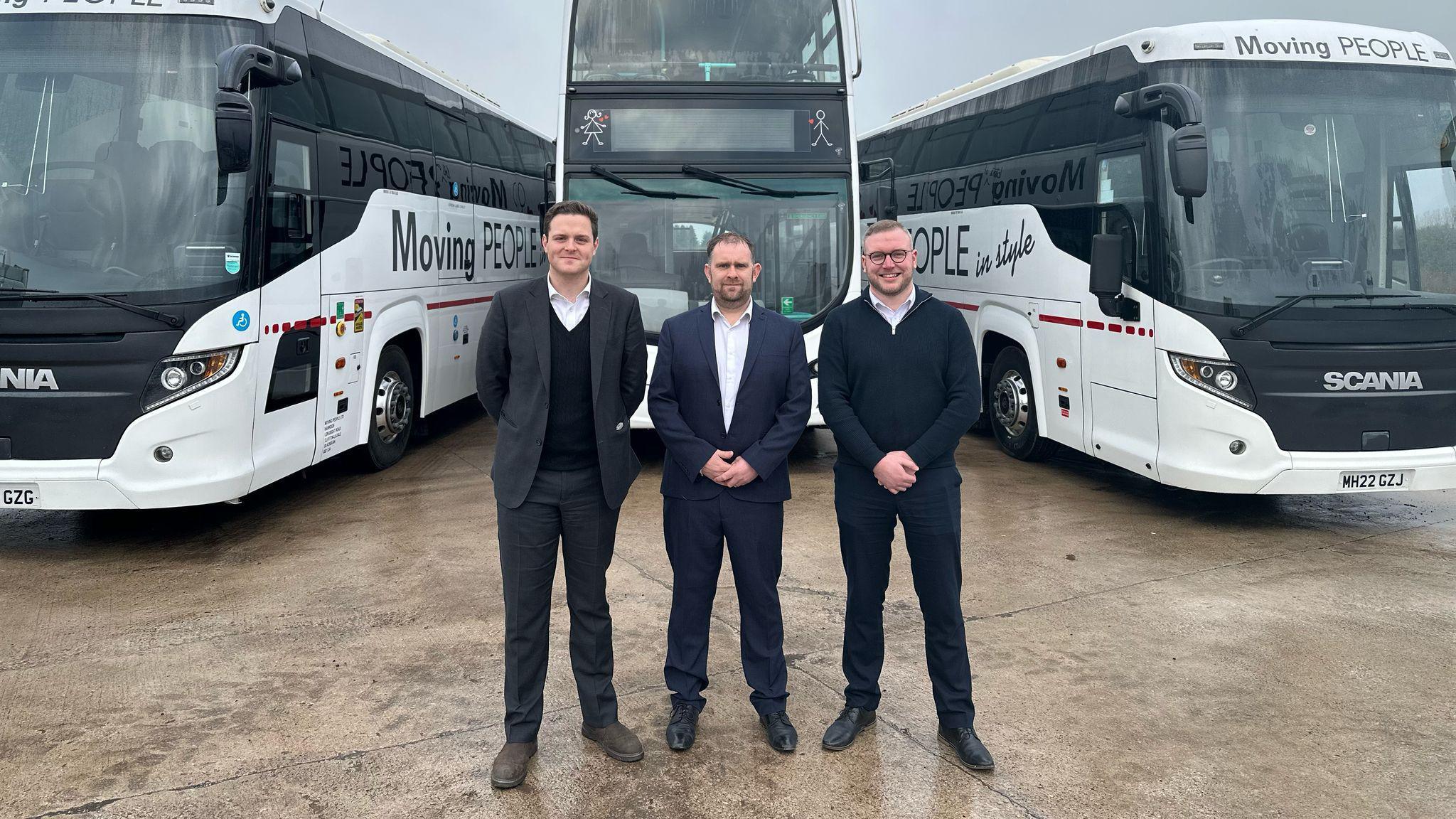 Three men stand in front of three white Moving People buses.
MP Oliver Ryan stands on the left, he has short hair, wears a dark suit, a white shirt and a black jumper. Moving People founder Stuart Coates stands in the middle, he has short hair, wears a dark suit and a white shirt. Councillor Scott Smith stands on the right, he wears dark trousers, a navy blue half zip jumper and a white shirt. 