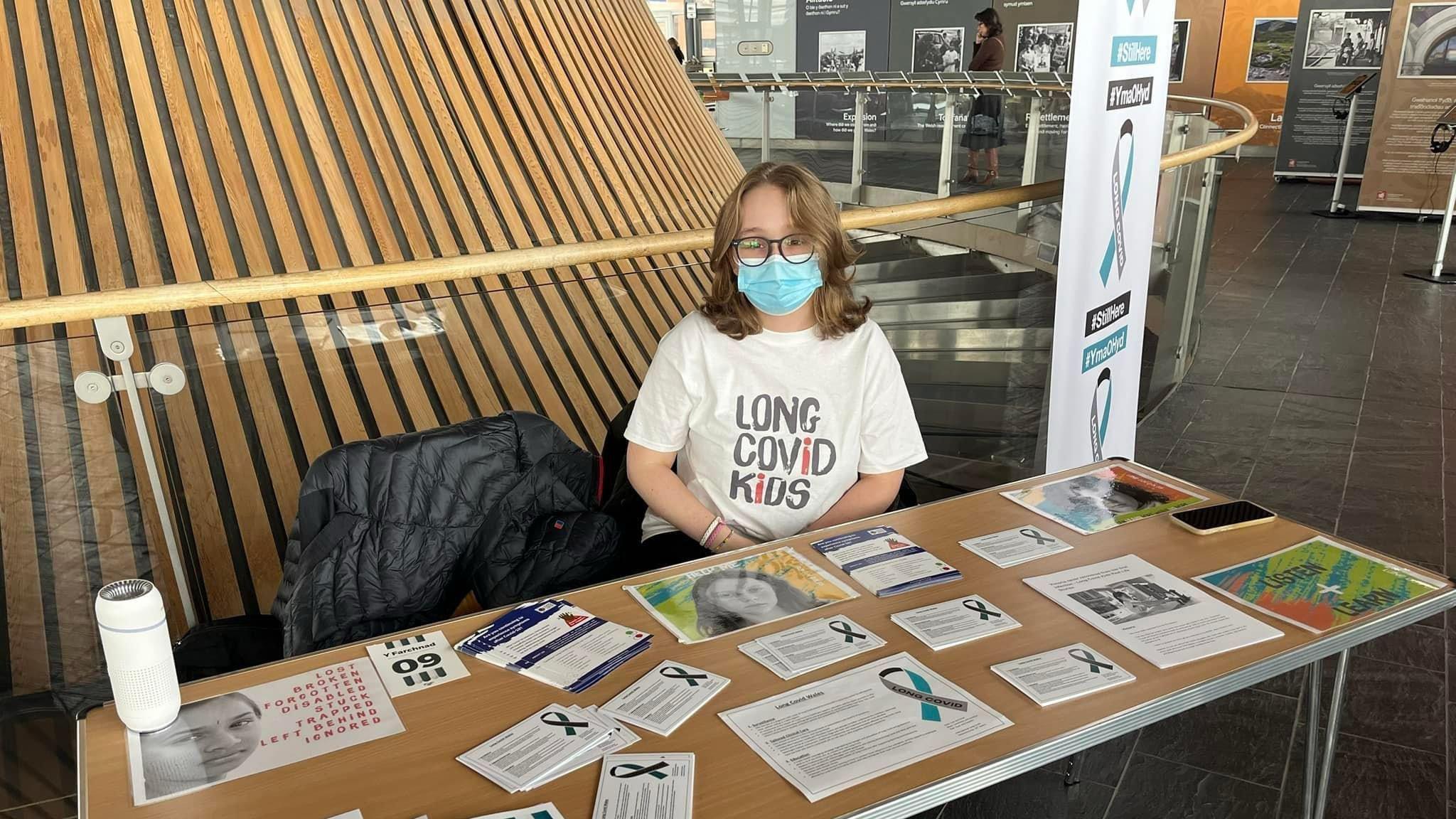 Yn y llun yma mae Bethan yn eistedd wrth ddesg yn Senedd Cymru yng Nghaerdydd. Ar y ddesg mae papuriau amrywiol sydd trafod profiadau plant a phobl ifanc o cofid hir.