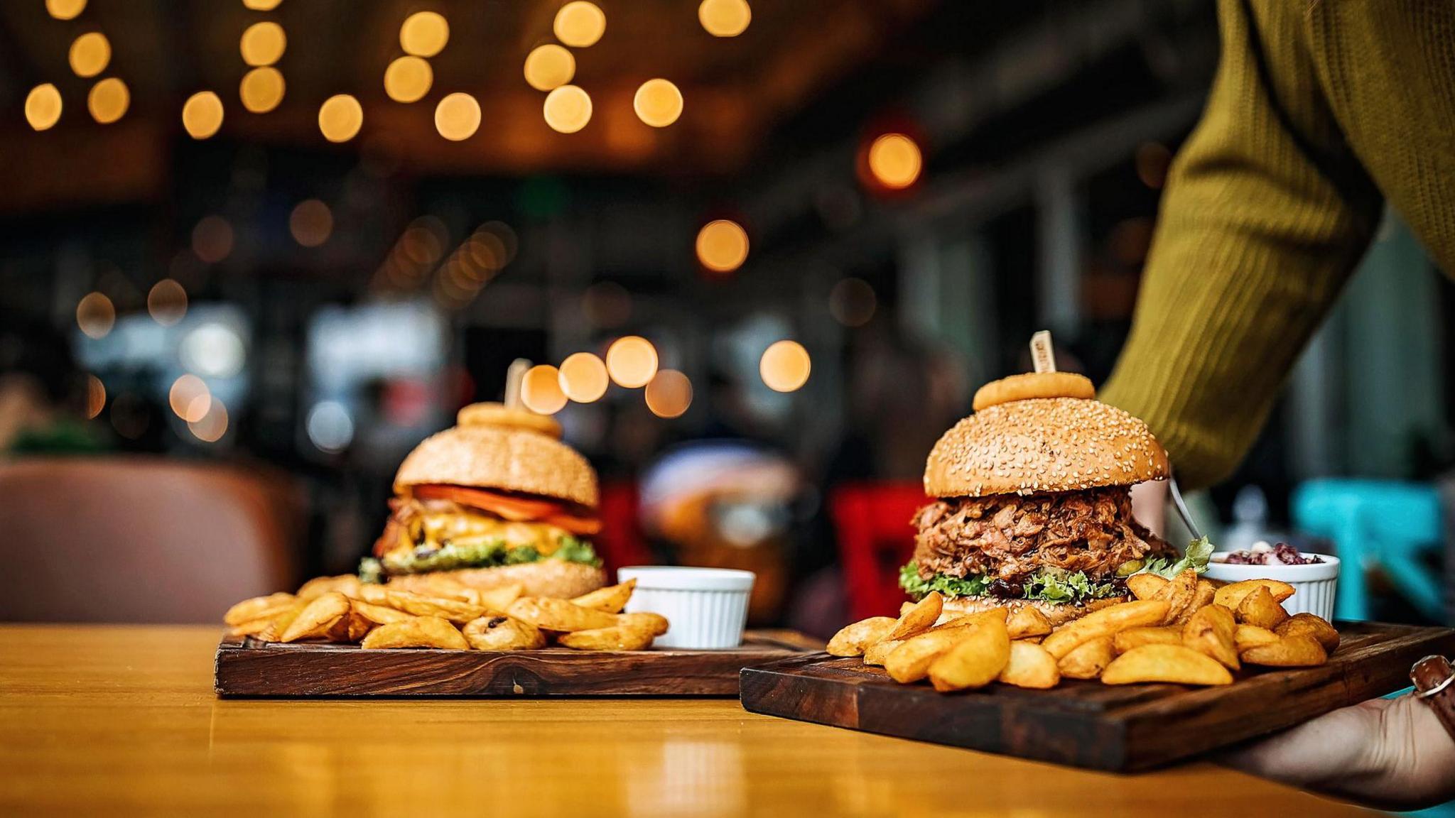 Two wooden boards, containing large burgers and chunky chips and sauces, are being delivered to a restaurant table in the evening, while lights are on in background