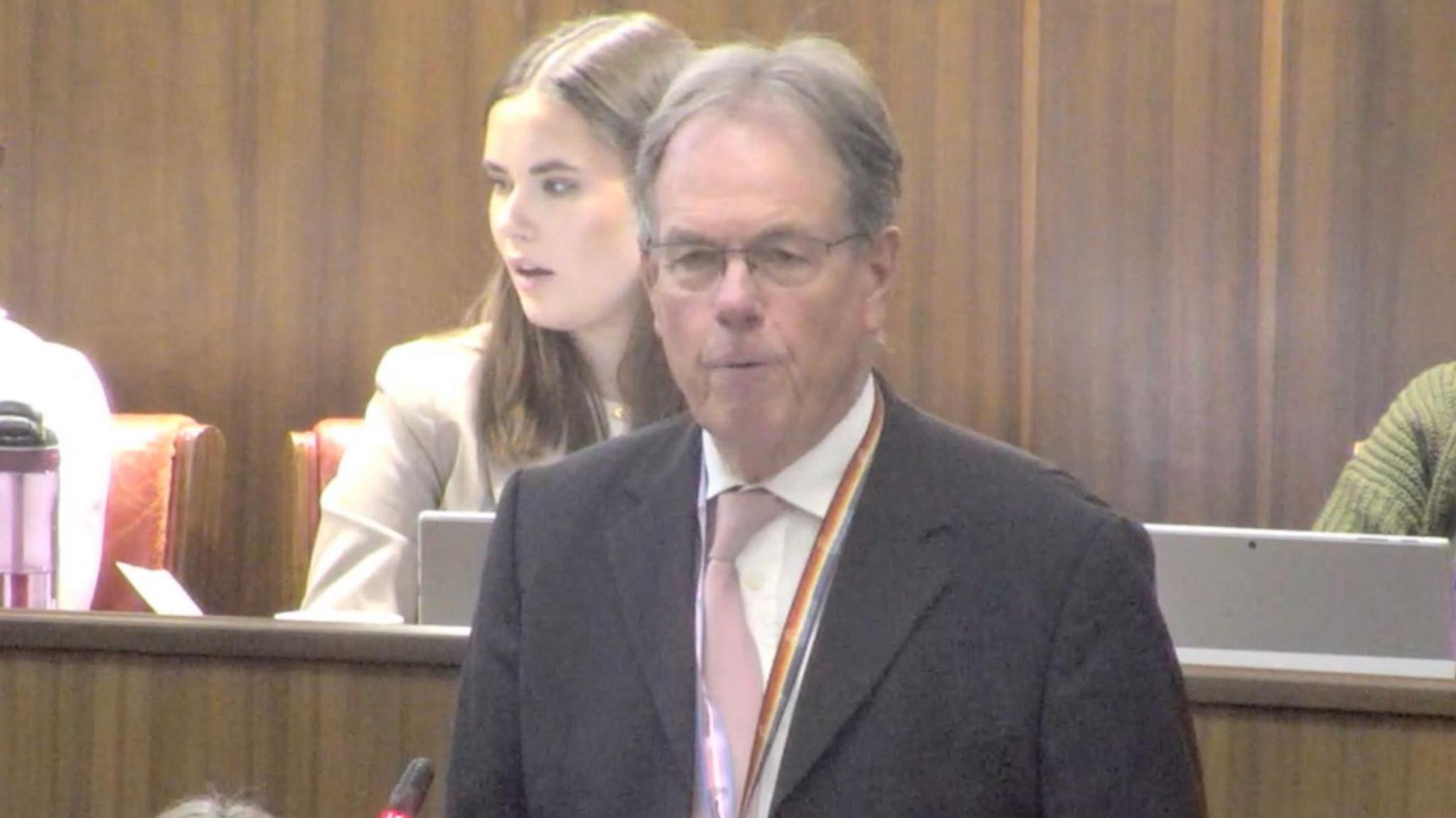 George Cowcher stands and speaks in the council chamber. Wood panelling is visible behind him. He wears glassed and a rainbow lanyard with a pink tie.