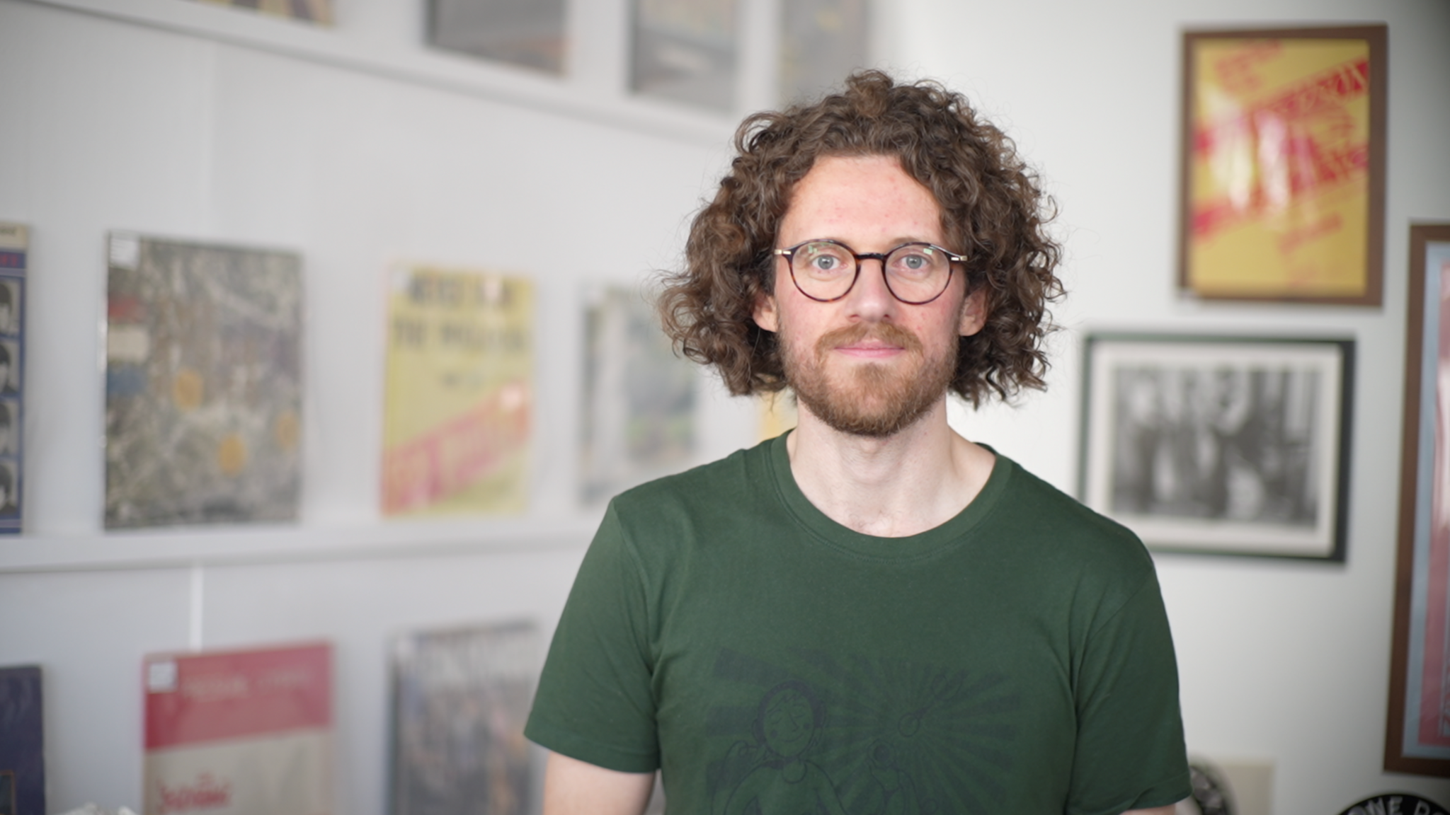 Frazer Merrick, standing in a record shop, with records behind him on the walls. He is looking straight at the camera, smiling and is wearing a green T-shirt. He has chin length curly dark hair, a beard and is wearing glasses.