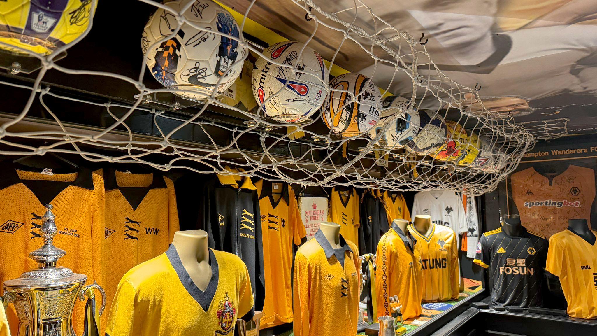 A collection of old gold and black Wolves football shirts in a museum and a collection of match footballs above on shelves