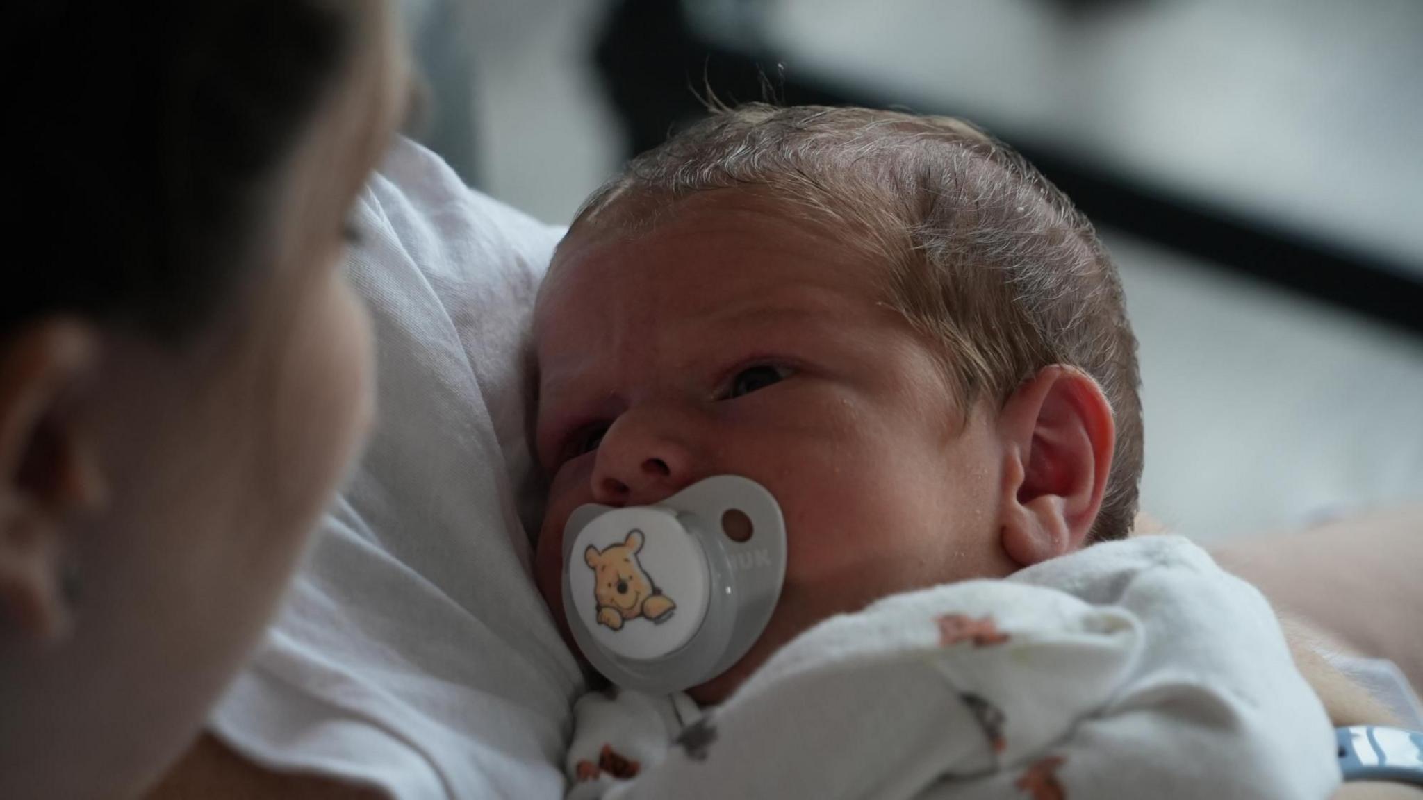 Mrs Robinson looks lovingly at her new son Henry Robinson. He is looking directly into her eyes. He still looks slightly wrinkled with a thin coating of brown hair. He has a Winnie the Pooh dummy in his mouth with a yellow bear in the middle of it and a white all in one.