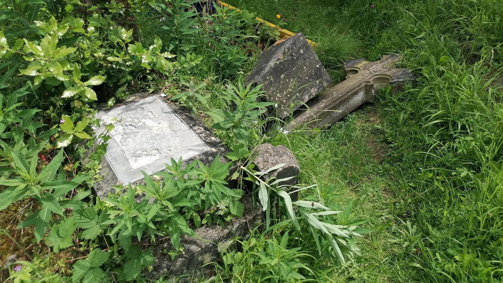 A cross statue which has been knocked off a stone plinth and lays discarded in the overgrown grass beside it.
