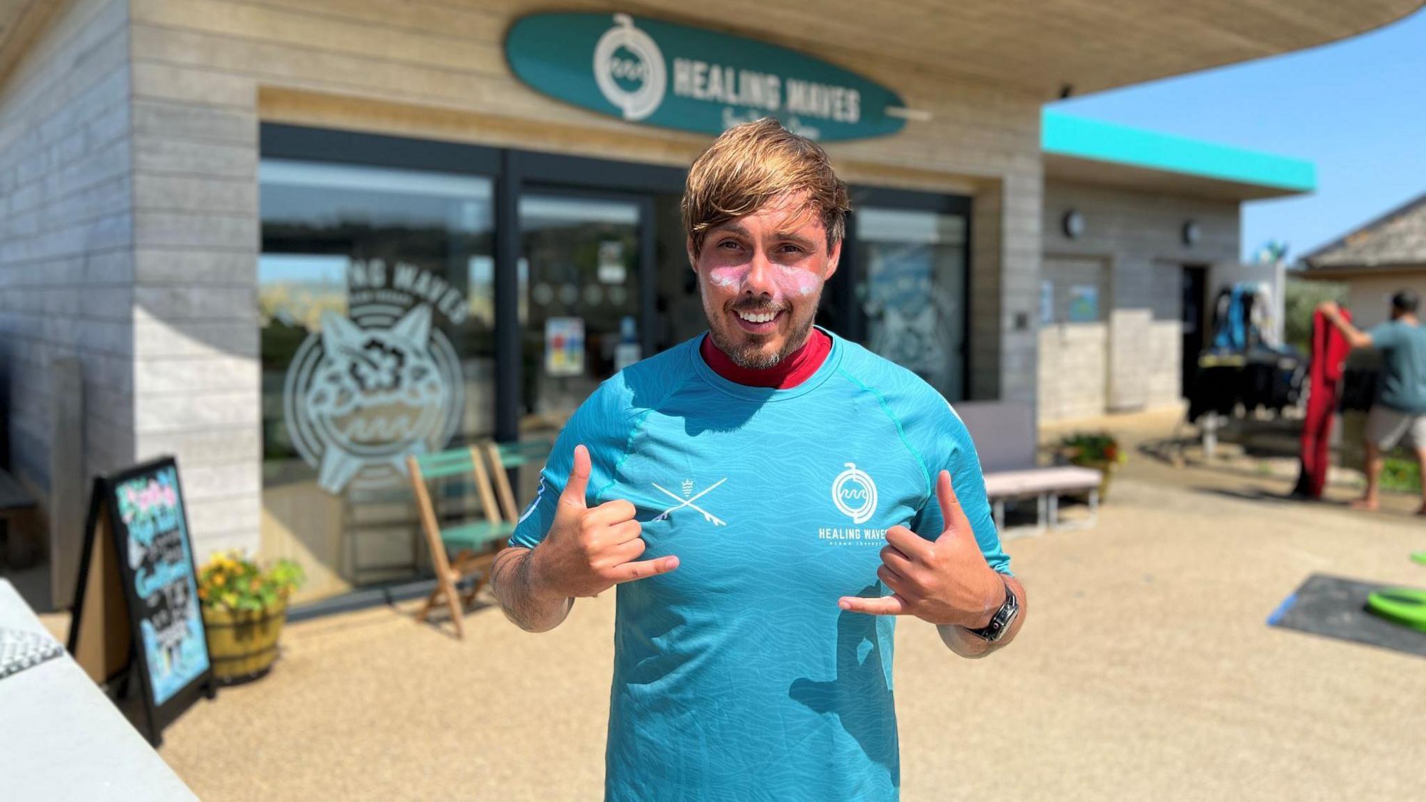 A man in a blue T-shirt with sun block on his cheeks smiles at the camera as he stands in front of the Healing Waves office - a modern, wooden structure with large windows near a beach.