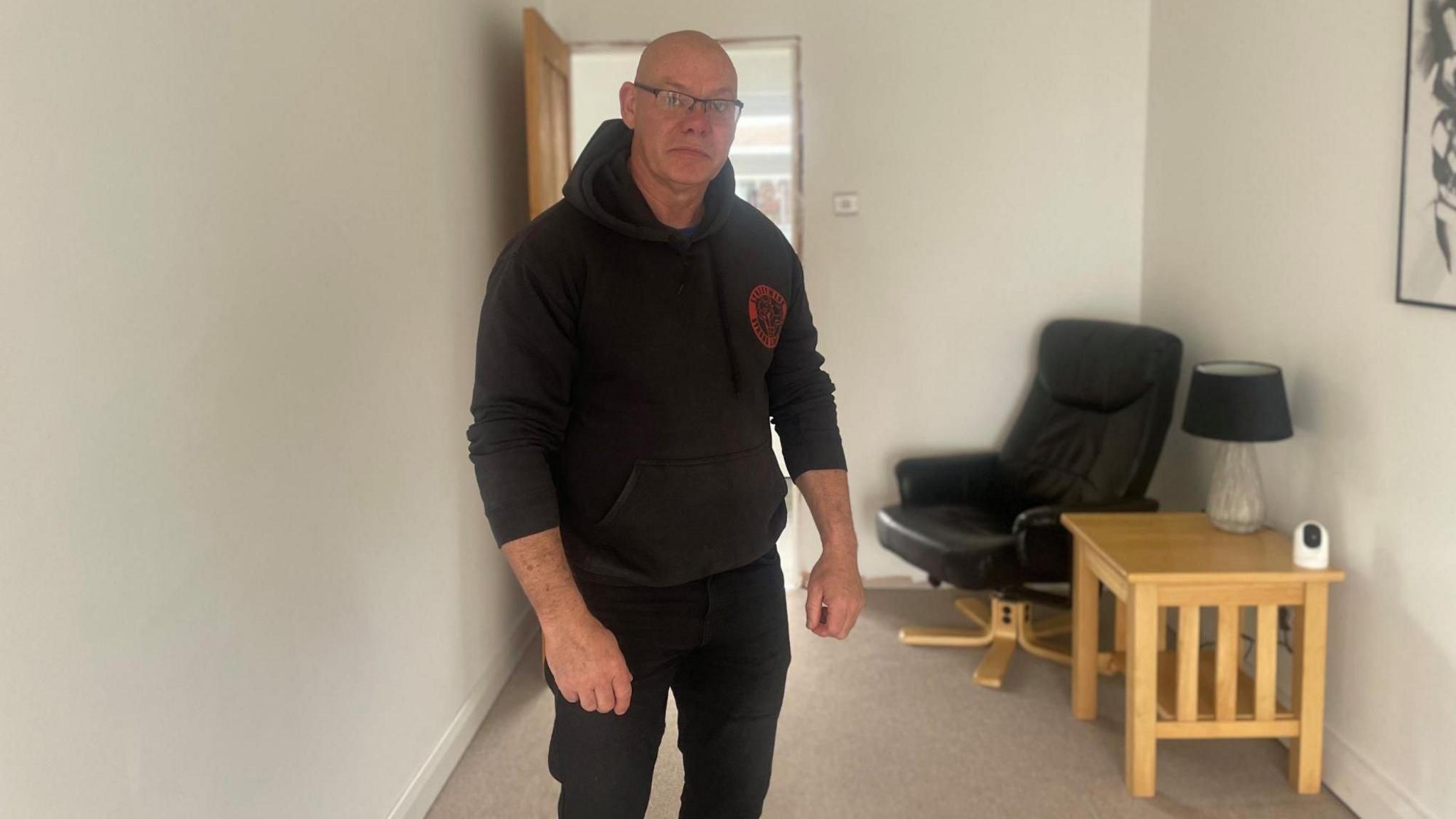 A bald man stands in a plain room with a beige carpet. He is dressed in a black hoodie and black jeans.