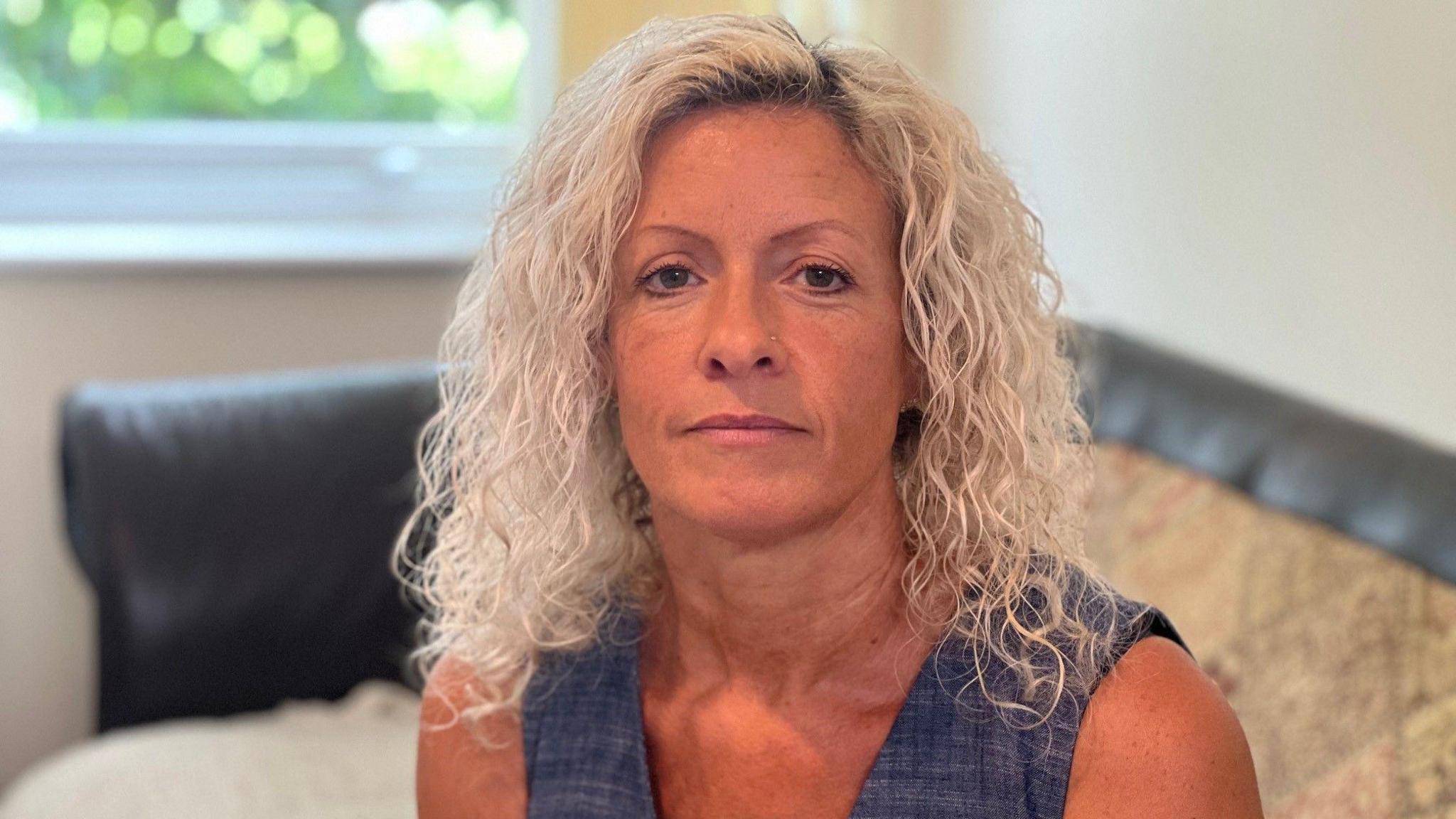 Michelle sat on a sofa looking at the camera. She has shoulder-length blonde curly hair, blue eyes and has a stud nose piercing. 
