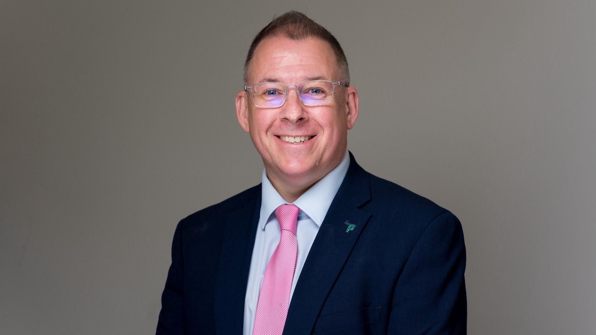 Professsional headhshot of Stephen McCarron. He is smiling into the camera wearing and suit and tie and glasses. He is in front of a grey background.