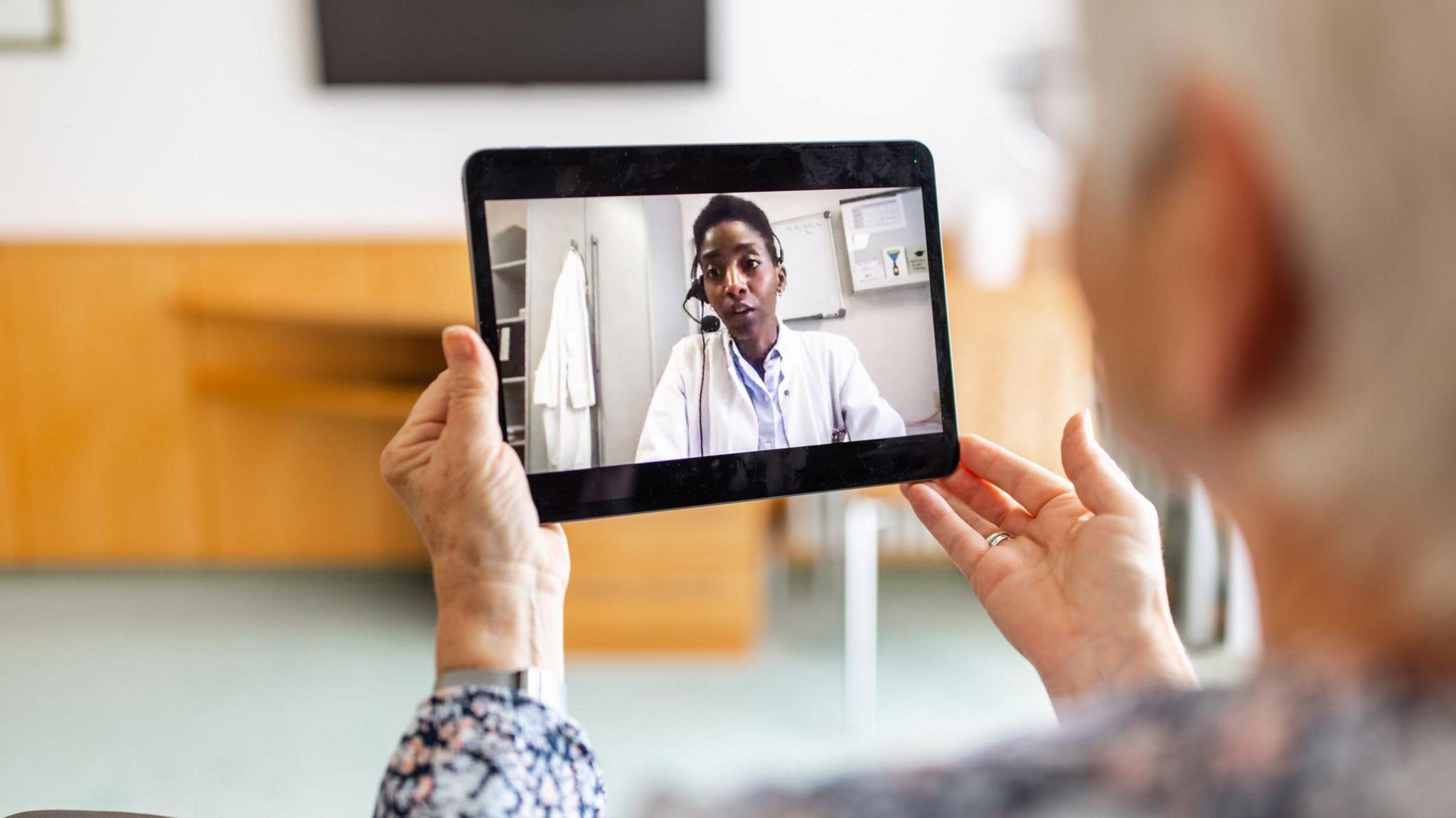 A blurred image of a woman in a living room holding up an electronic tablet. On the screen is an image of a woman - wearing a white medical coat and a headset - talking to her.