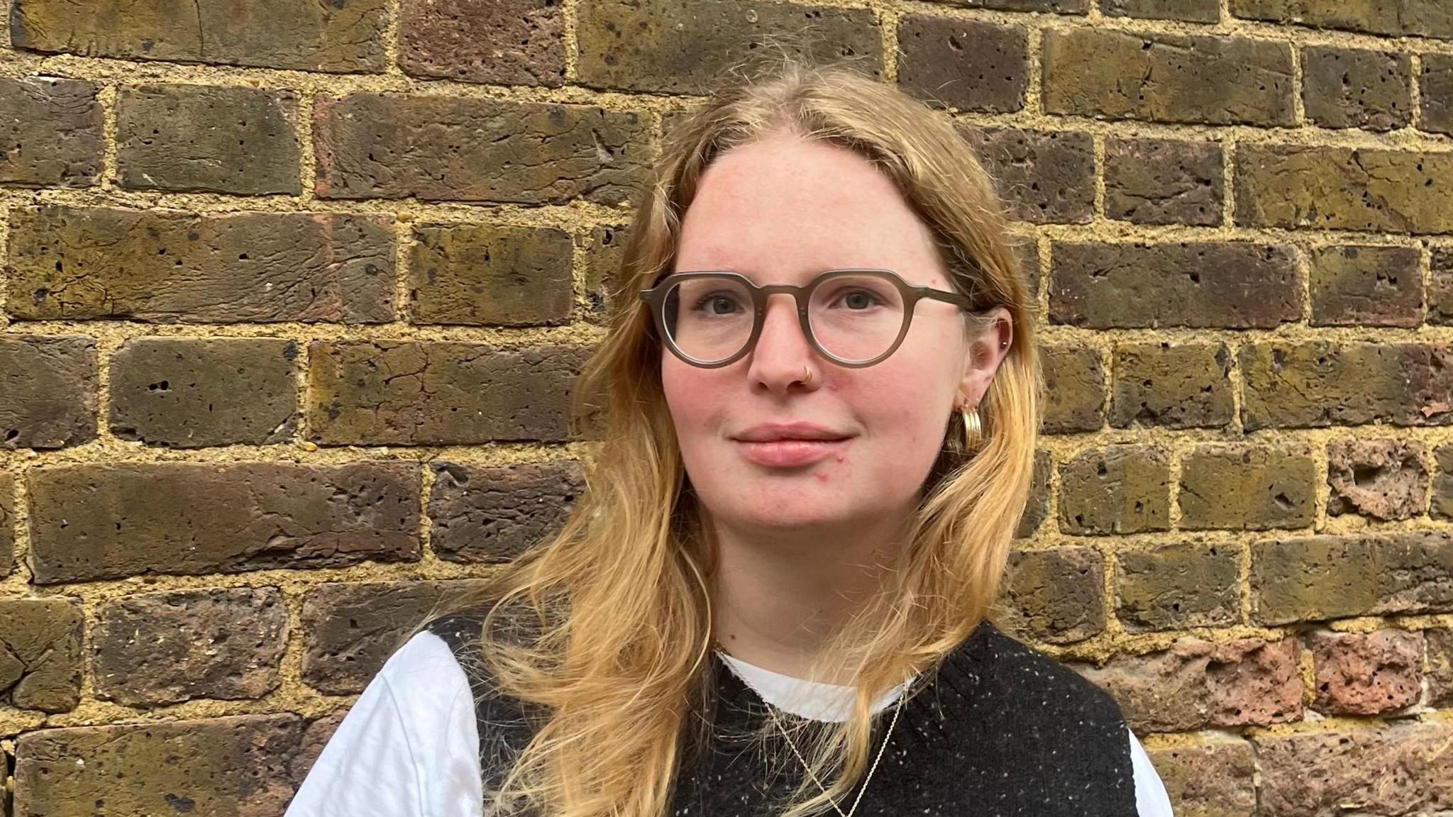 Hattie wears glasses and has blonde hair. She is wearing a black sweater vest with a white t shirt underneath.