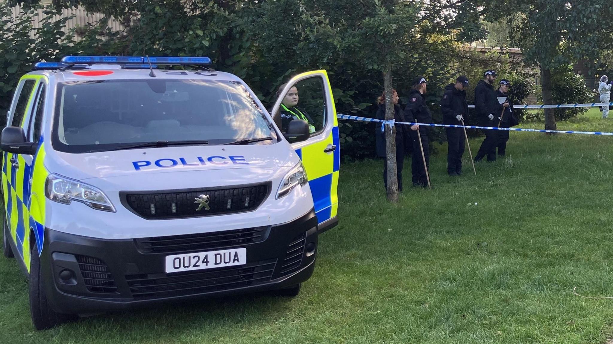 Police car parked on grass and uniformed officers, one standing by the open door of the car, five others behind blue and white police tape. A forensic officer can just be glimpsed in white jumpsuit in the distance on the right.