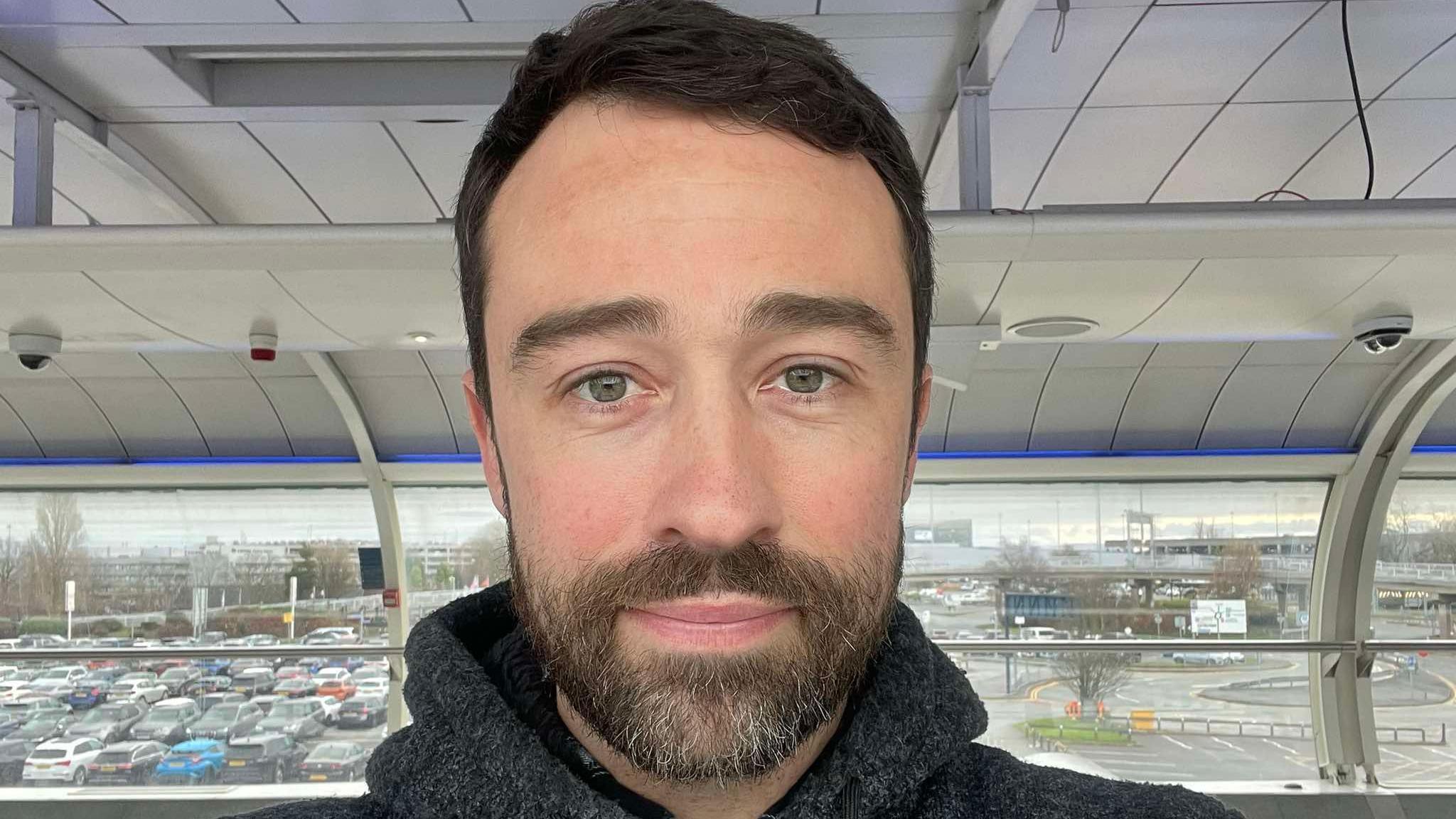 A bearded man looking at the camera with an airport scene in the background