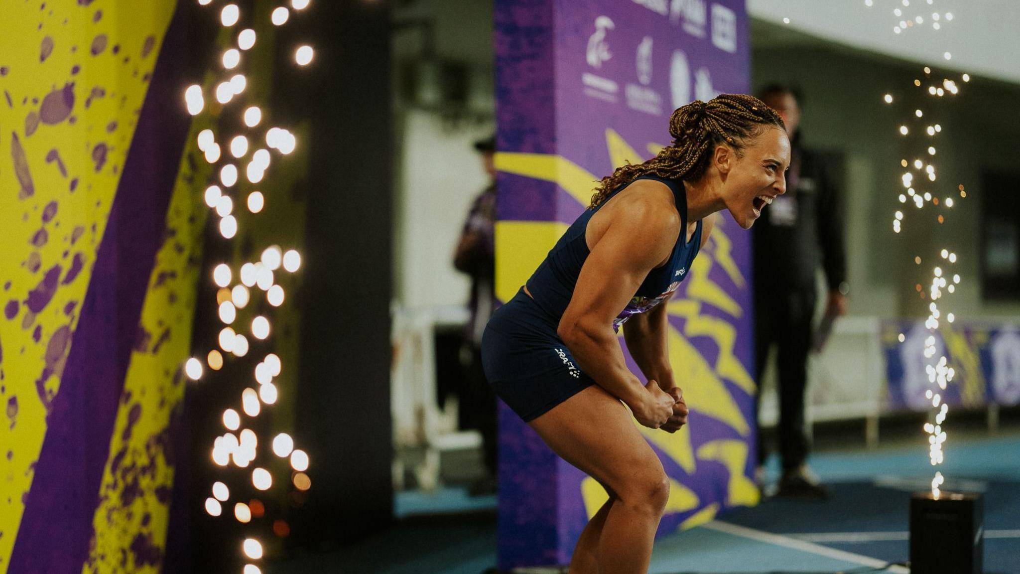An athlete is on a stage and looks to be celebrating. There are pyrotechnics at either side of the front of the stage. The female athlete is posing with a big smile. 