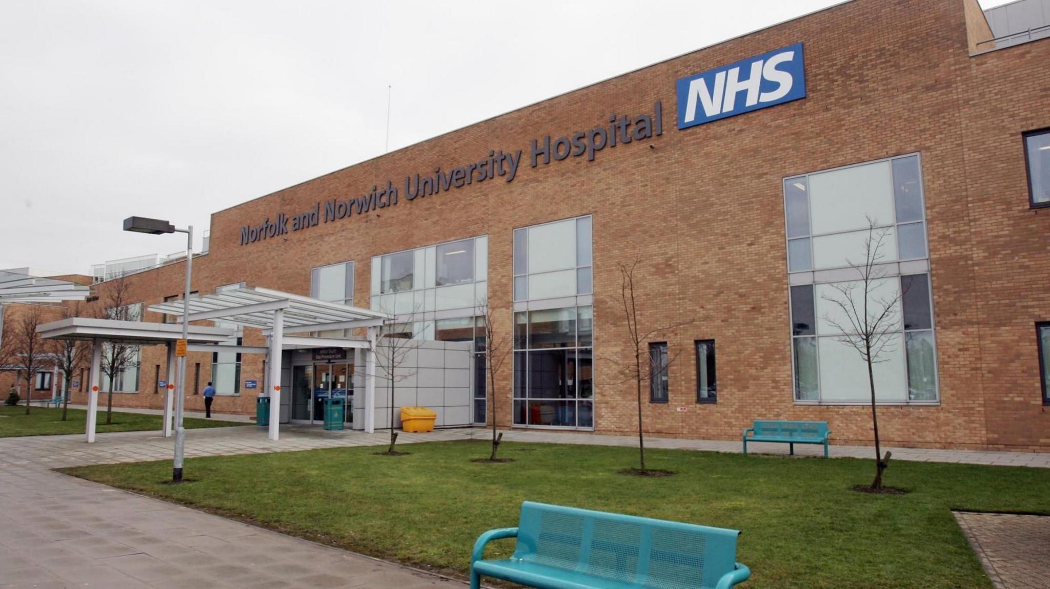 The outside of the Norfolk and Norwich University Hospital where the laboratory for the diabetes test for Norfolk is. The picture shows the front entrance with the name of the hospital and NHS in bold letters on the side of the building. The building is made of tan coloured brick with large windows. There is a grassed area to the front