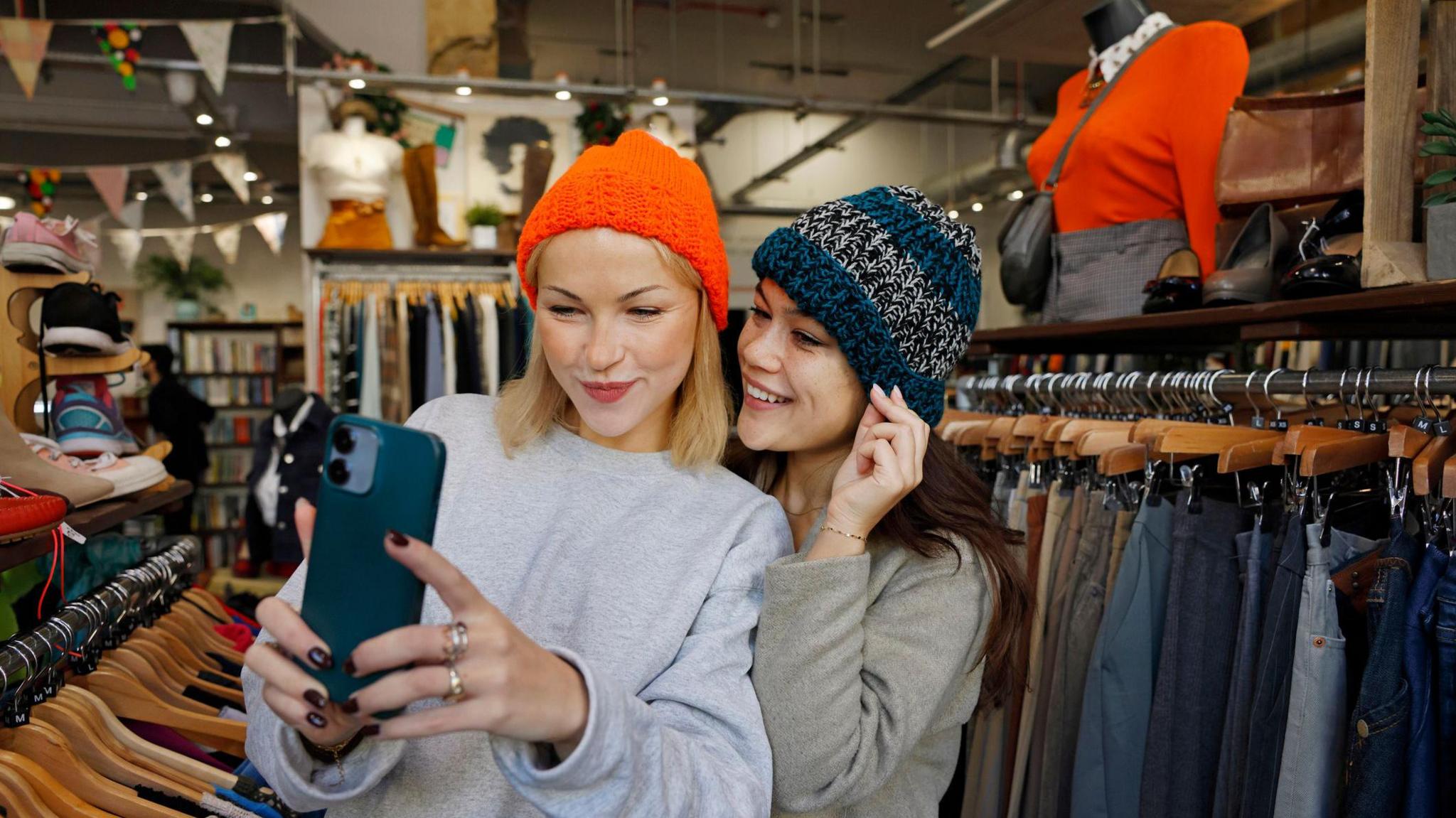 Two female shoppers take a selfie while trying on woolen hats in a clothes store