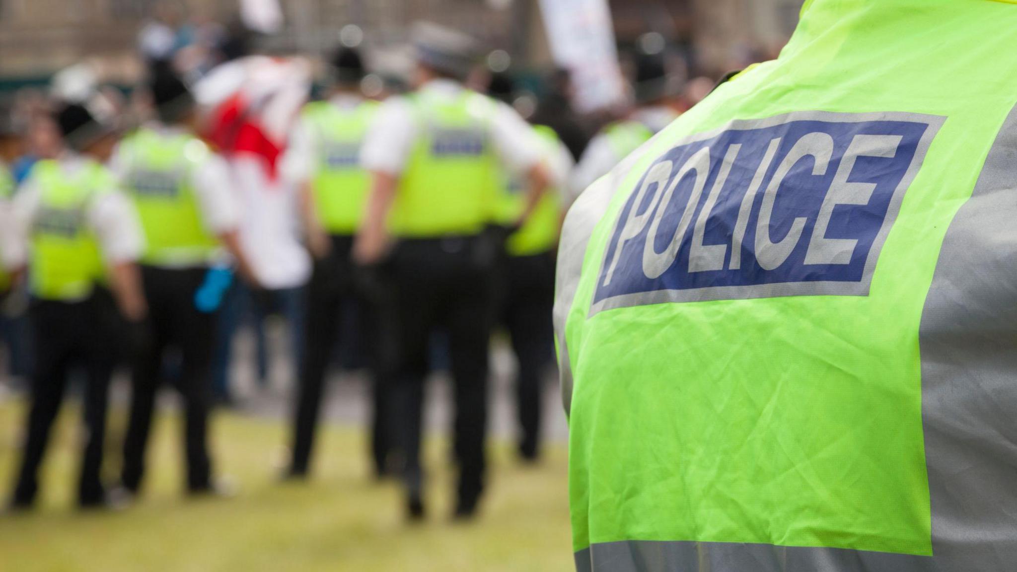 A picture of the back of a police officer wearing a green jacket with "police" on the back of it, with five others further away from the camera. They are not in focus and are blurred.