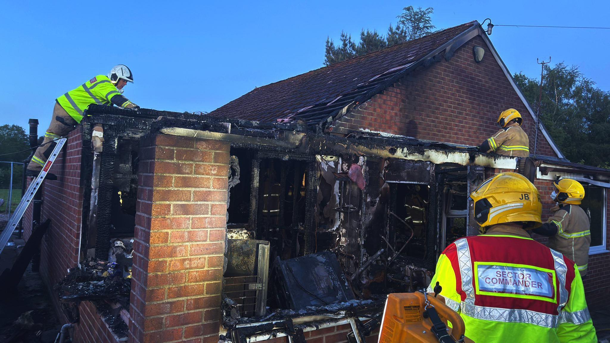 Firefighters work at the scene of a blackened, burnt-out bungalow. A room is exposed and the insides are charred. One of the firefighters is on a ladder.