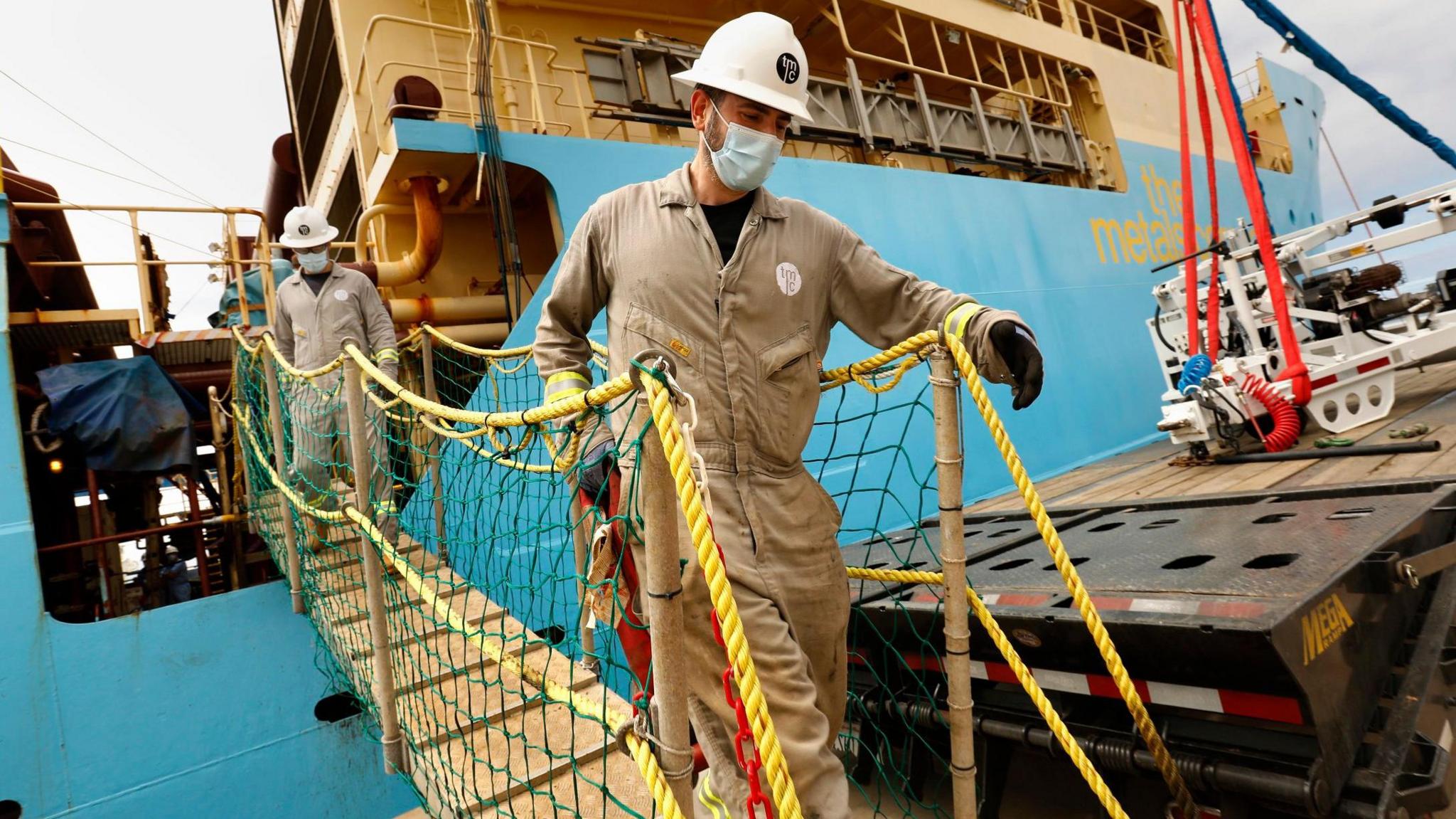 Two men dressed in brown jumpsuits and white hard hats walk down a gangway from a blue ship