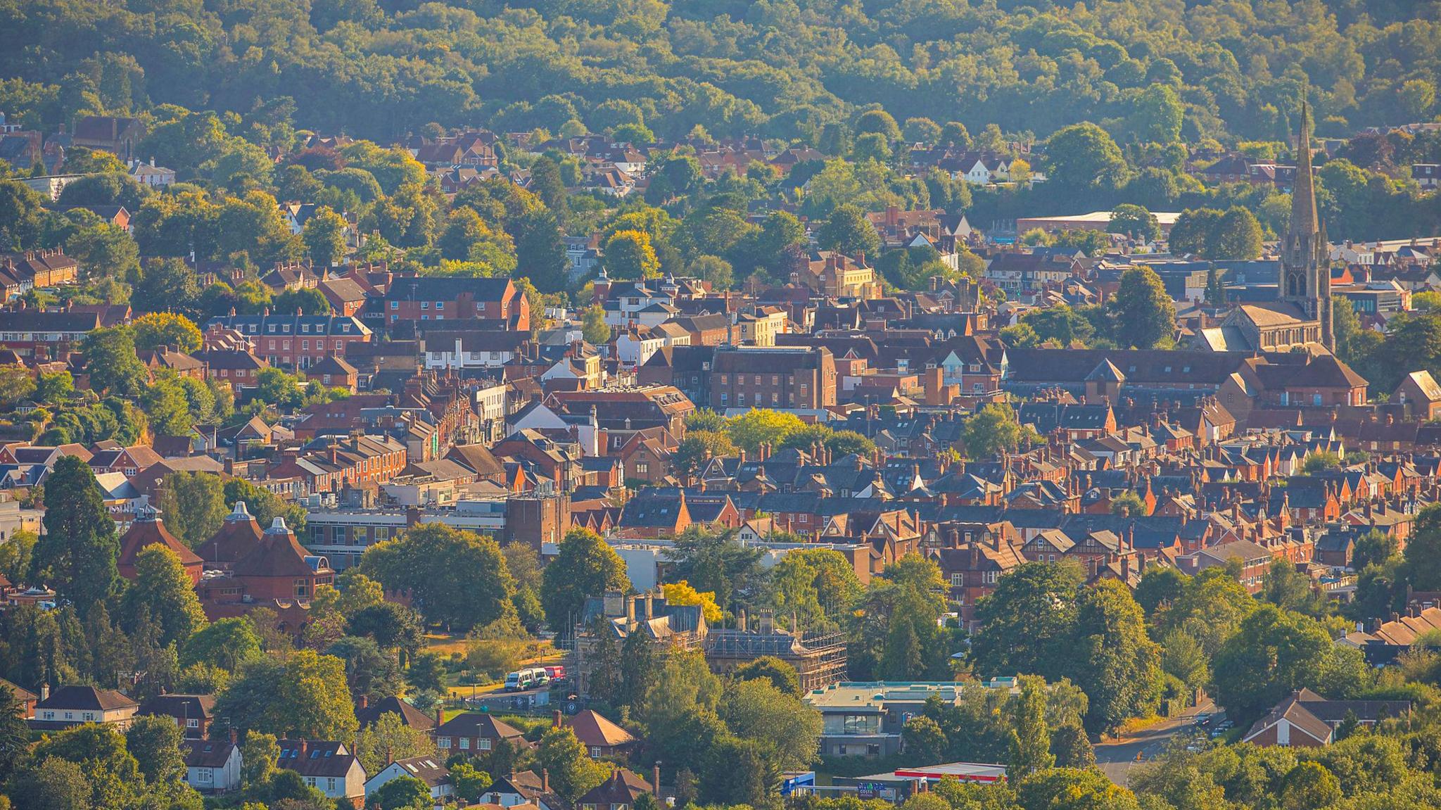 Rows of houses are seen from the distance in a town that slopes gently uphill. There is a church spire to the right of the shot and the sun is shining on the homes and surrounding trees.