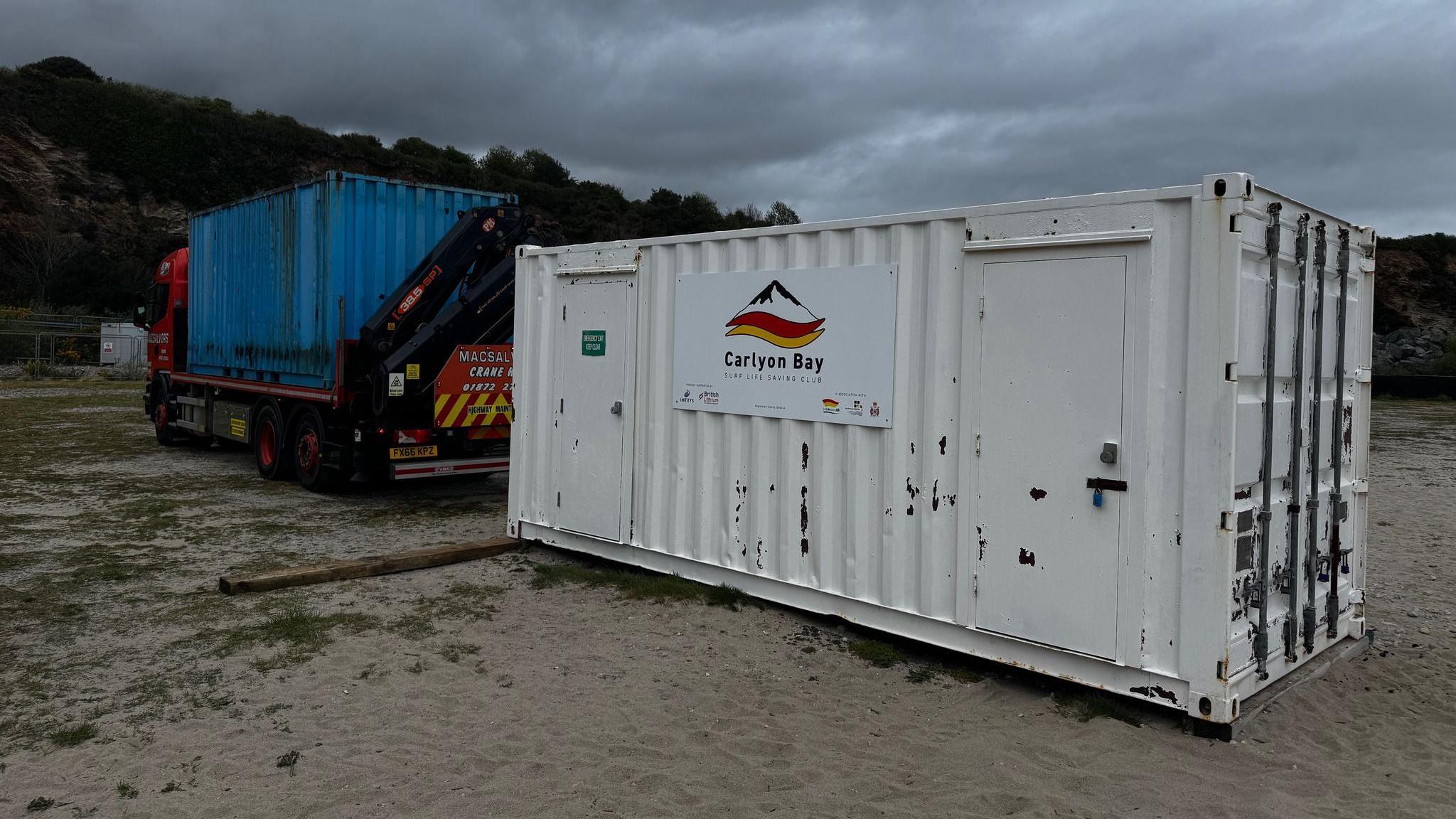 A white shipping container with a sign attached to it. There is a lorry with a blue container on the back.