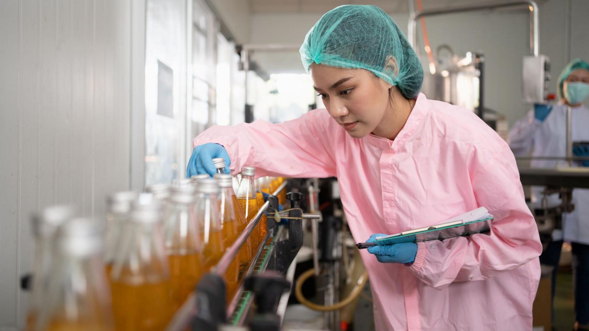 A woman in a blue hair net wearing a pink coat and blue gloves inspects a glass bottle on a factory production line