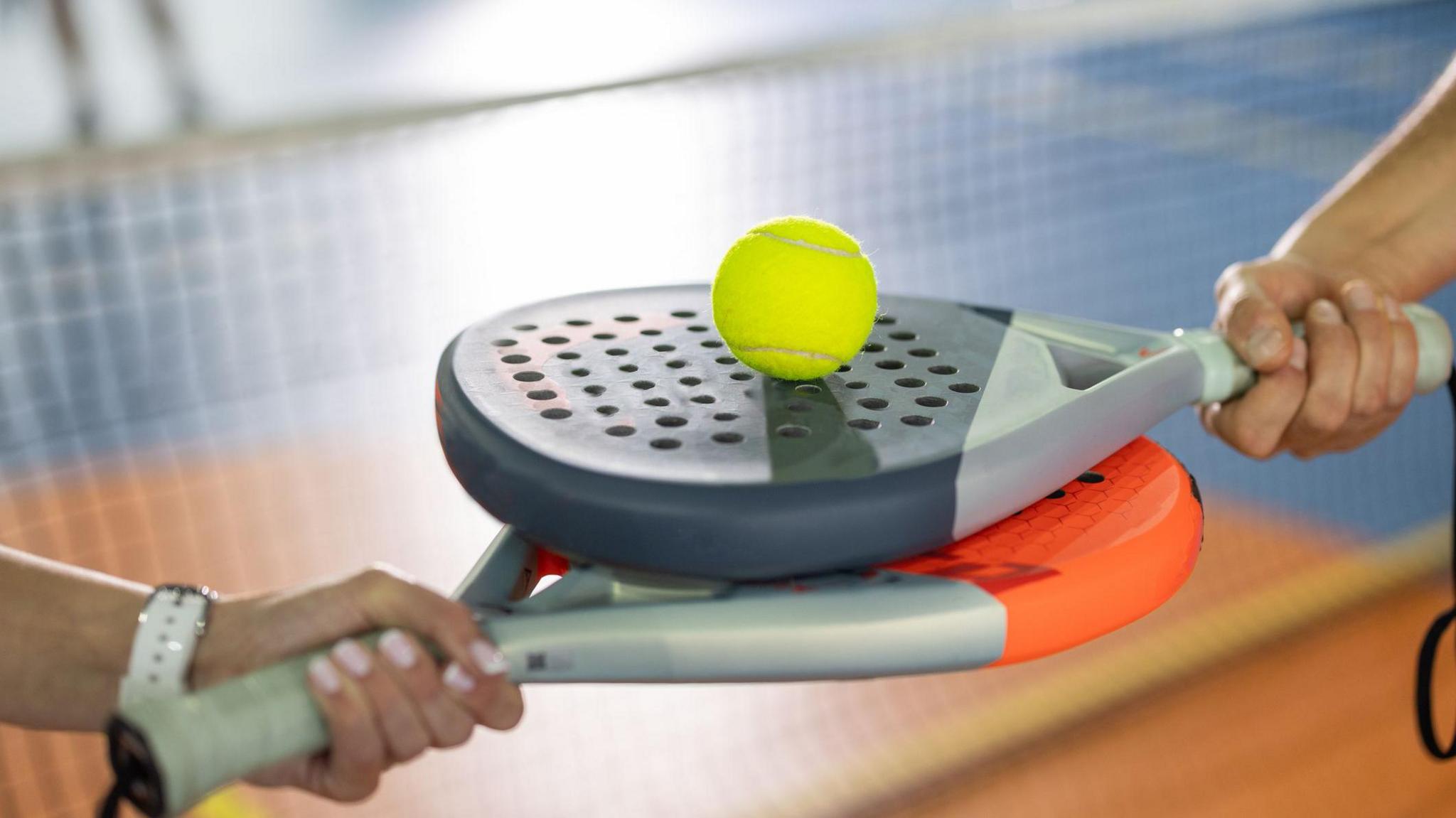 Two people holding padel racquets horizontally, with a padel ball sitting on top. There is a padel net blurred in the background.