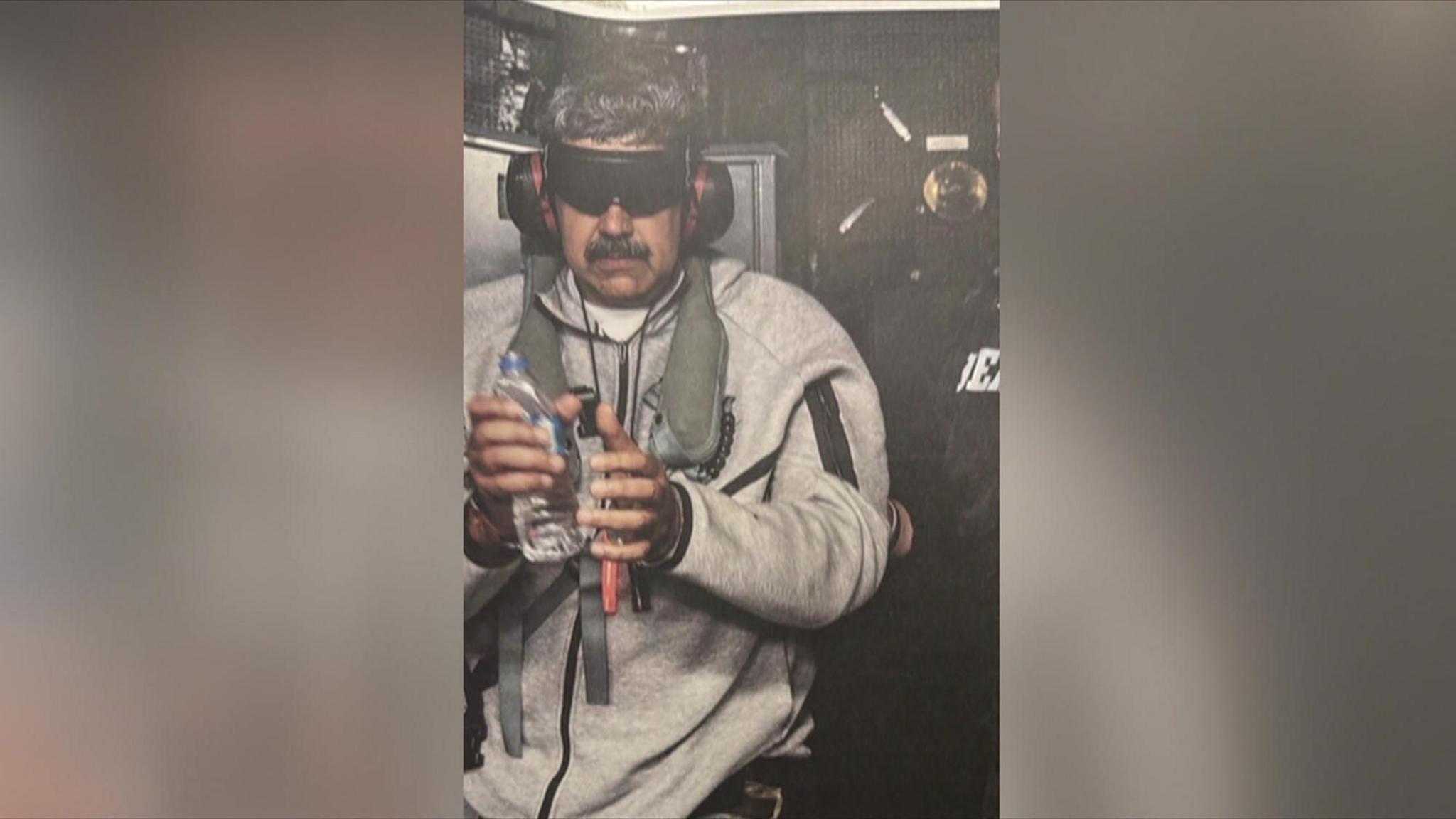 Maduro on the USS Iwo Jima, wearing a grey tracksuit and holding a bottle of water. He is blindfolded and wearing ear protection.