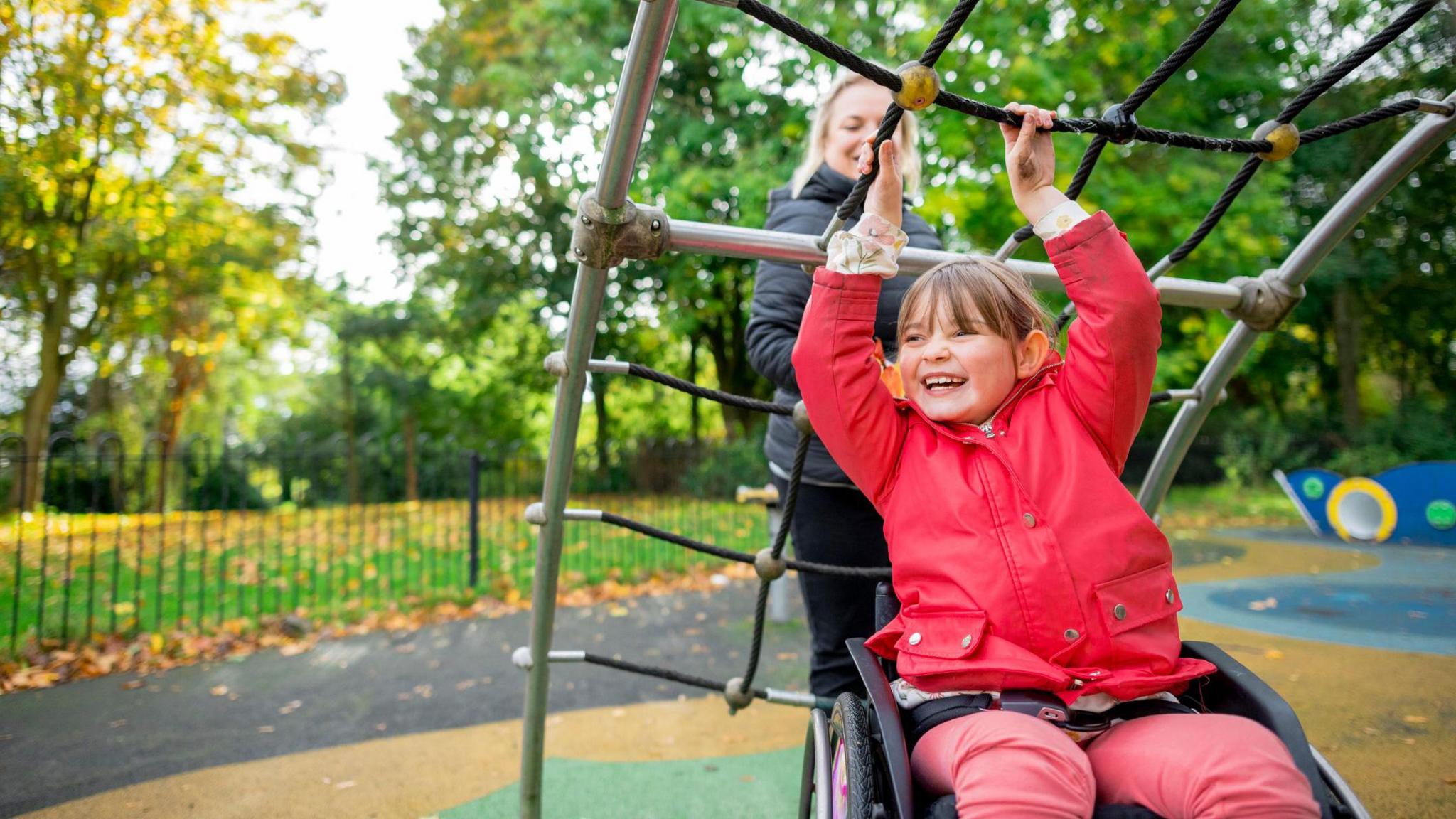 A girl using a wheelchair and dressed in a red rain mac holds onto a rope bridge in a playground with her mum behind her