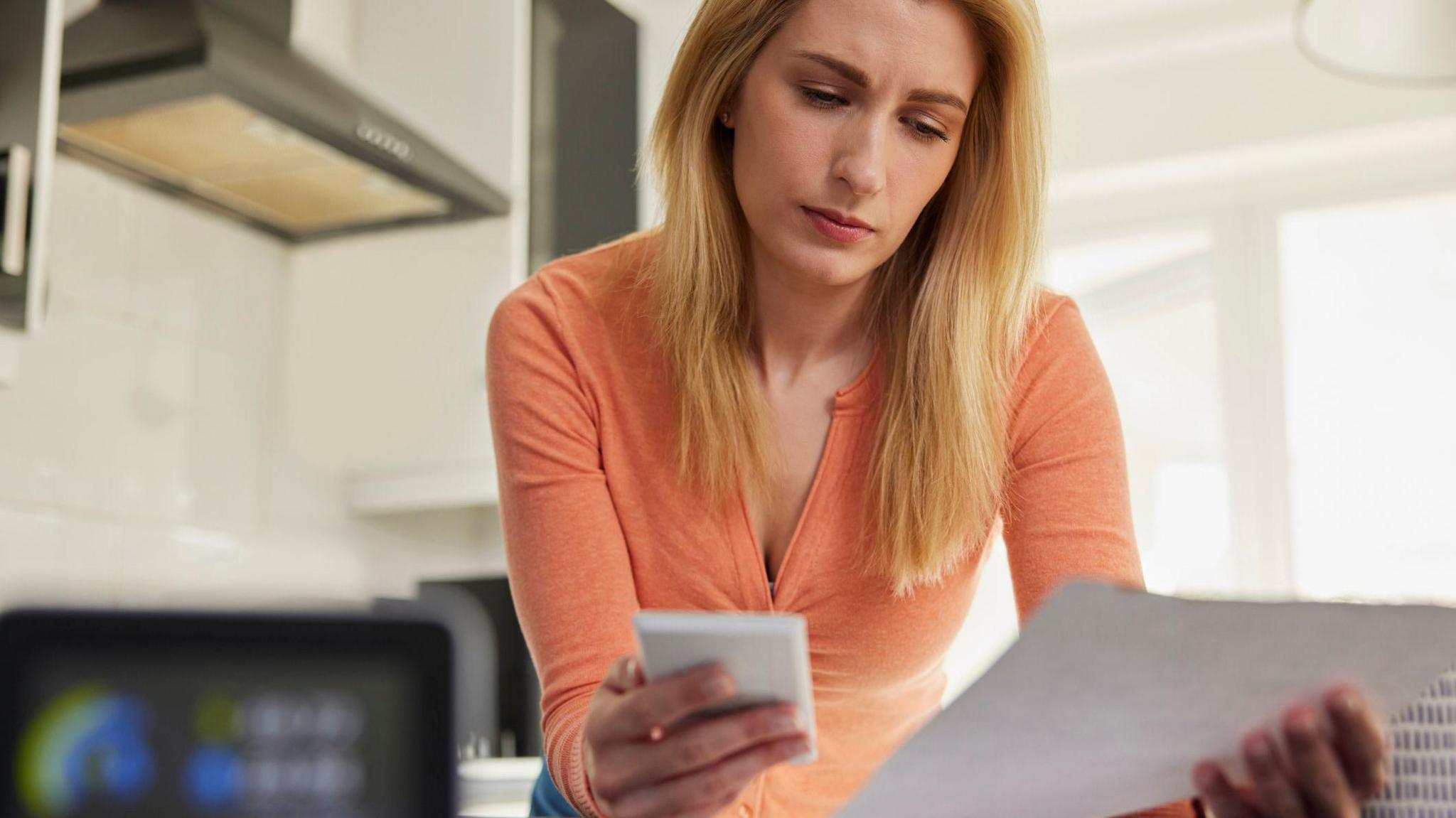 Blonde woman in a kitchen, looking at her phone and a bill at the same time.