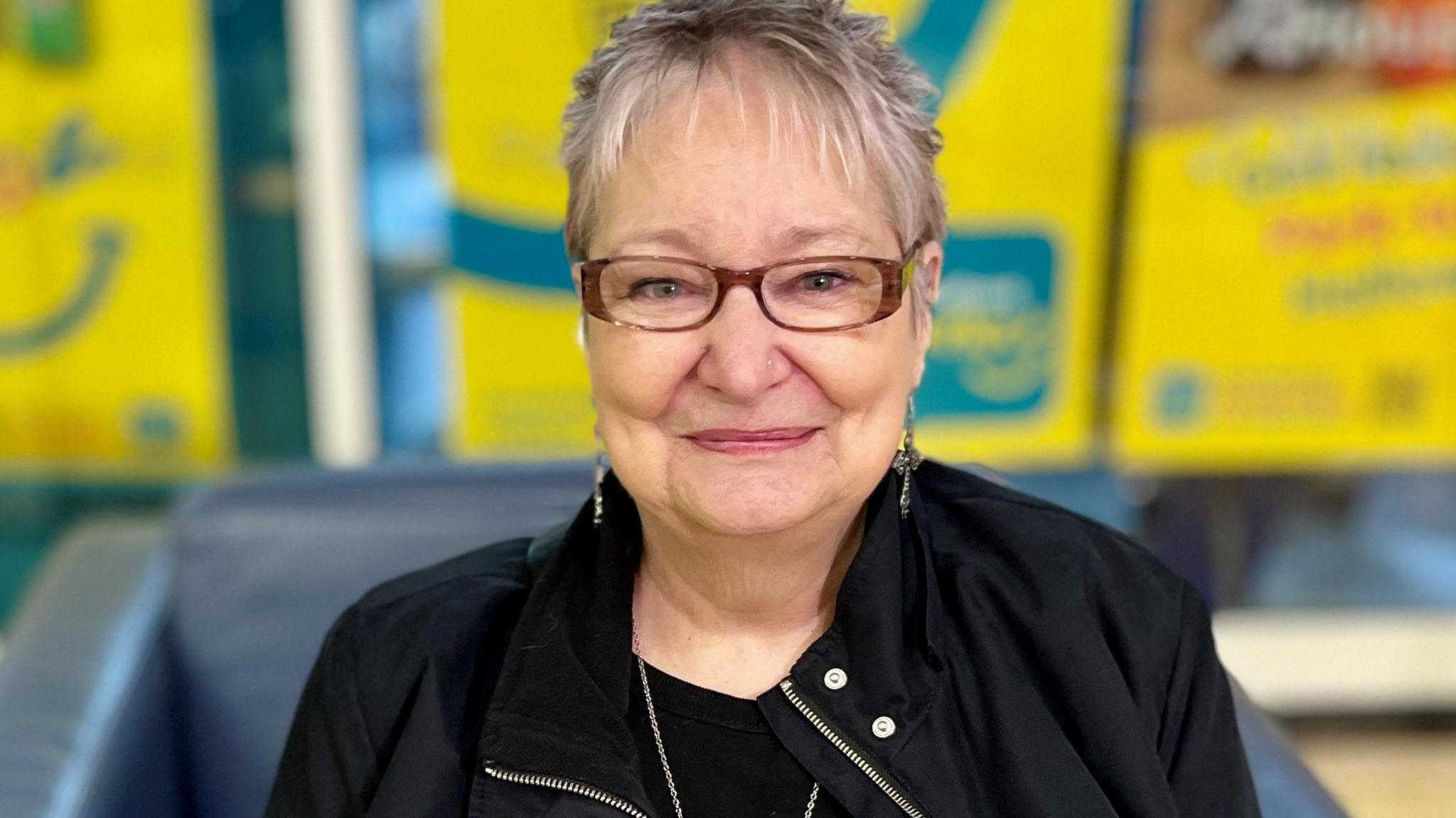 Liz has short grey hair with a feathery fringe and is wearing dark brown framed glasses and dainty drop earrings and a black jacket over a black top. She is smiling into the camera as she sits in front of a shop window