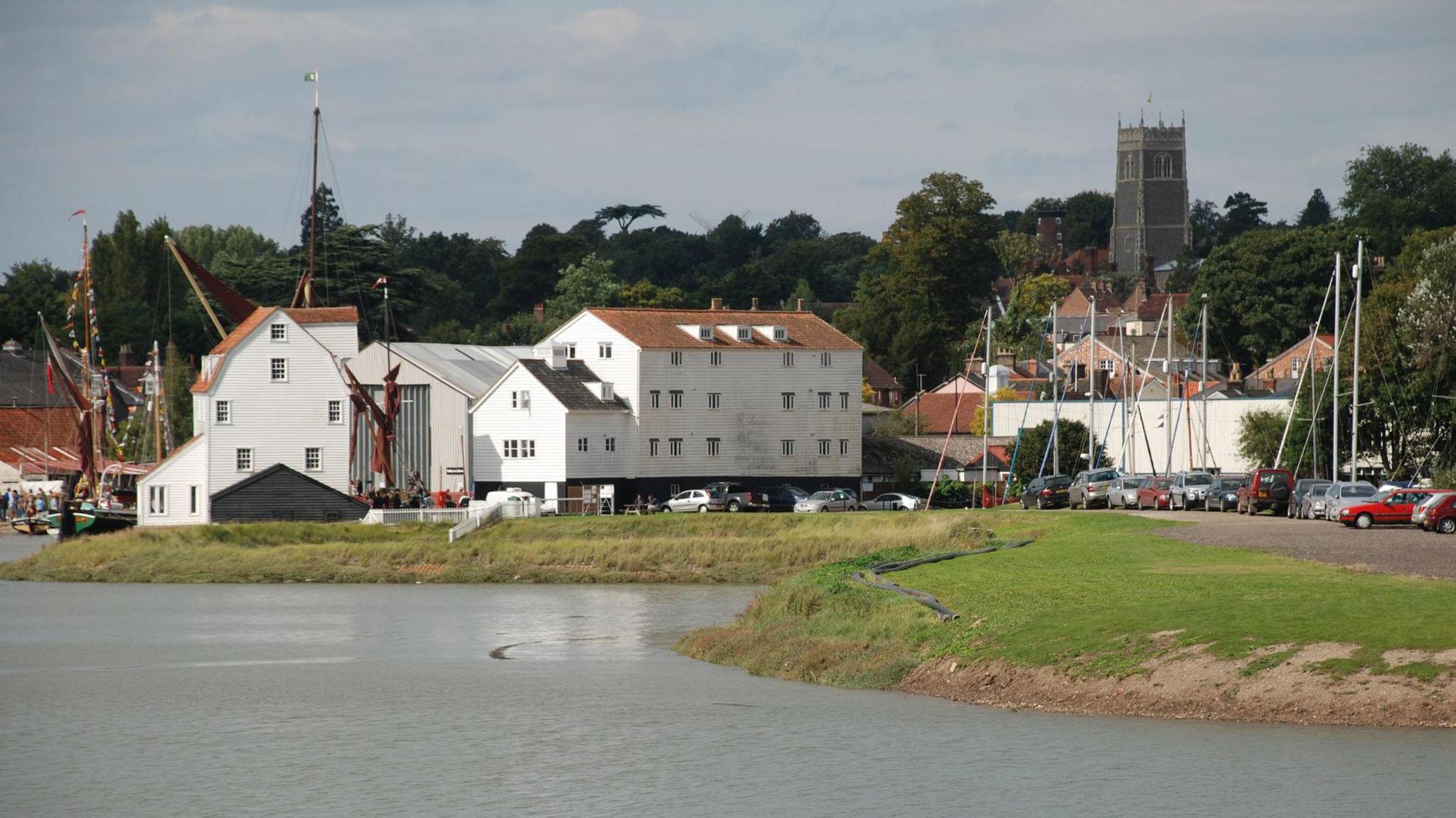 The tide mill, houses, cars and a church spire are all visible near the banks of the River Deben in Woodbridge. Boat masts are visible and the river is positioned in the foreground.