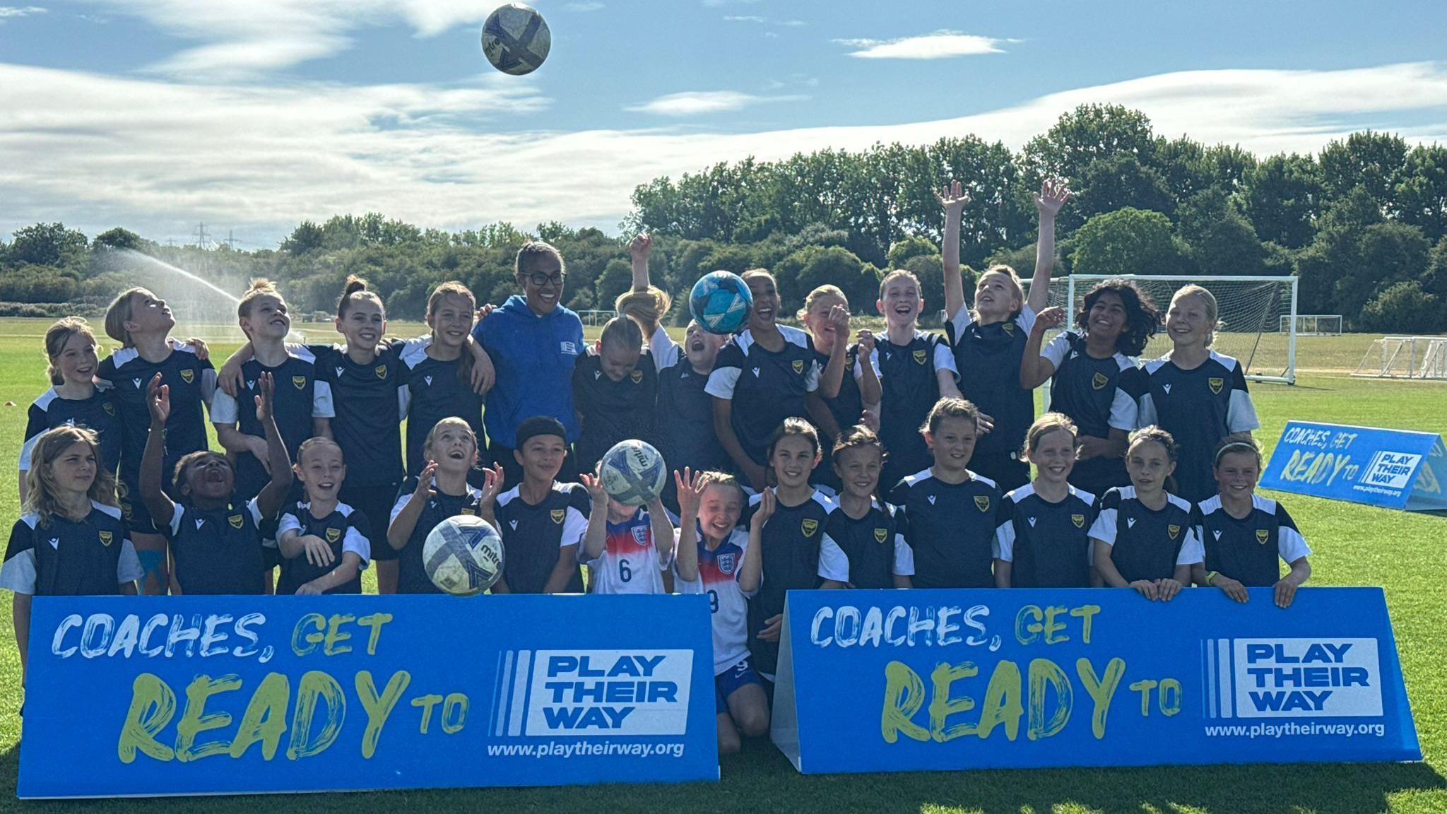 A group picture of smiling Rachel Yankey with the girl's team that she visited. Some of the girls are looking at footballs thrown in the air. It is sunny.
