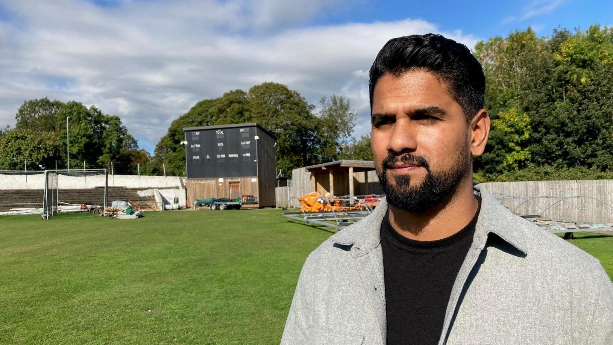 A man wearing a grey shirt and black T-shirt underneath standing in front of scoreboard on a green cricket pitch