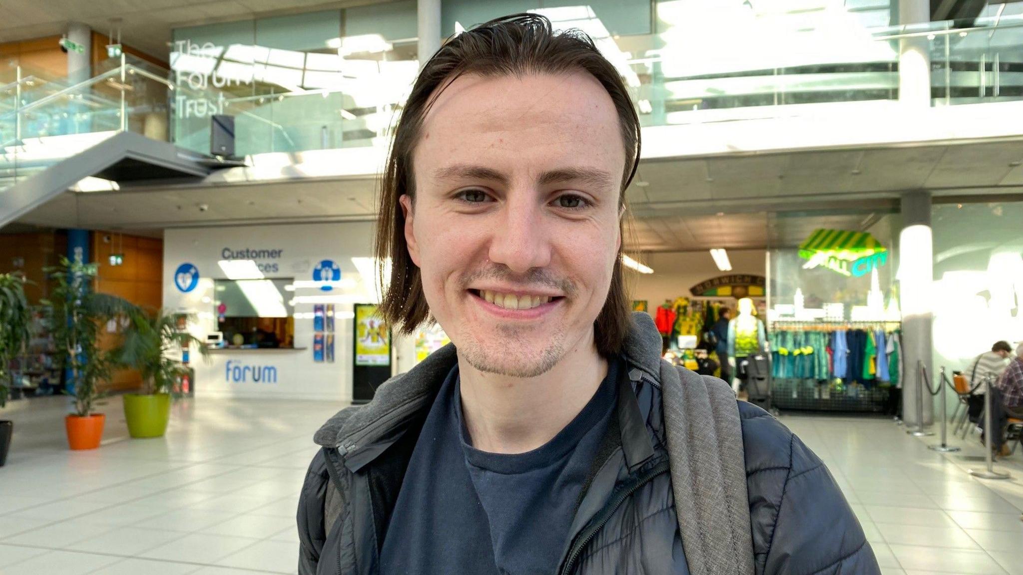 Young man smiling and wearing dark T-shirt and jacket. There are shops behind him and three pot plants.