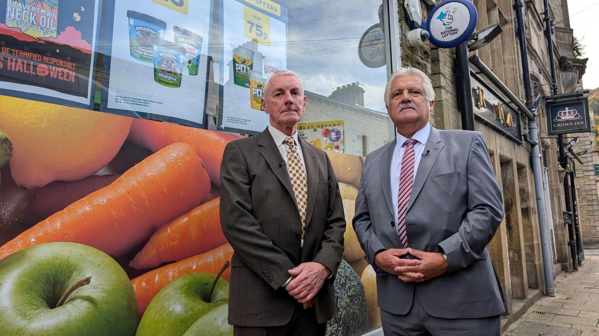 A picture of retired police officers Tony Whittle and Graham Sunderland. They are both wearing suits and are stood outside a shop on a street in Hebden Bridge. 