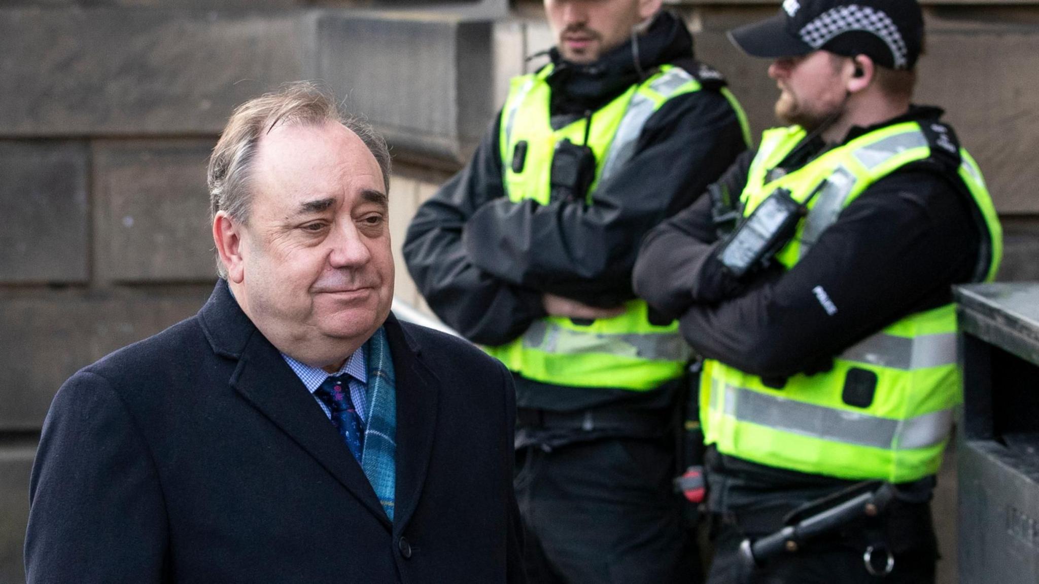 Alex Salmond walks past two police officers outside the High Court in Edinburgh during his trial. He is wearing a dark blue coat and a tartan scarf