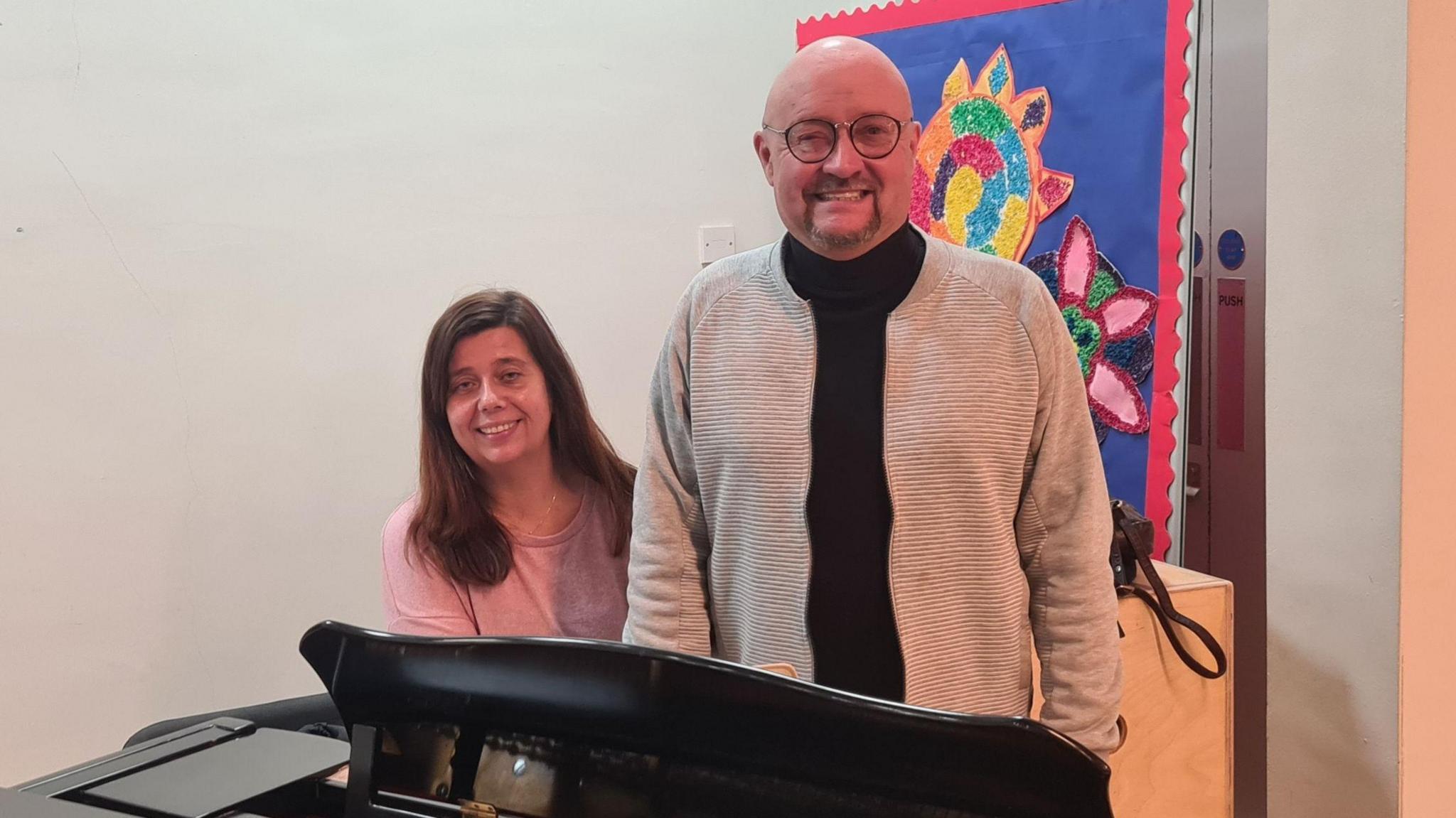 Ryan Longridge and Sarah-Jane Cooper by the communal grand piano in 2 Royal Avenue.  Sarah-Jane has long, dark hair and is wearing a pink top.  Ryan is bald with a goatee beard.  He is wearing glasses, a black top and beige cardigan.