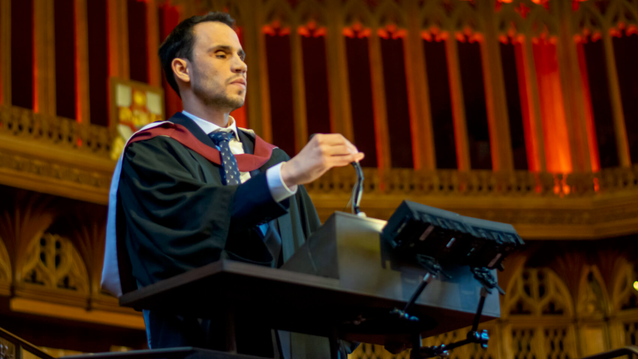 Mr Fattouh is photographed wearing a black robe with red and white accents, as well as a blue tie. He is standing in front of a dark brown plinth with a microphone.