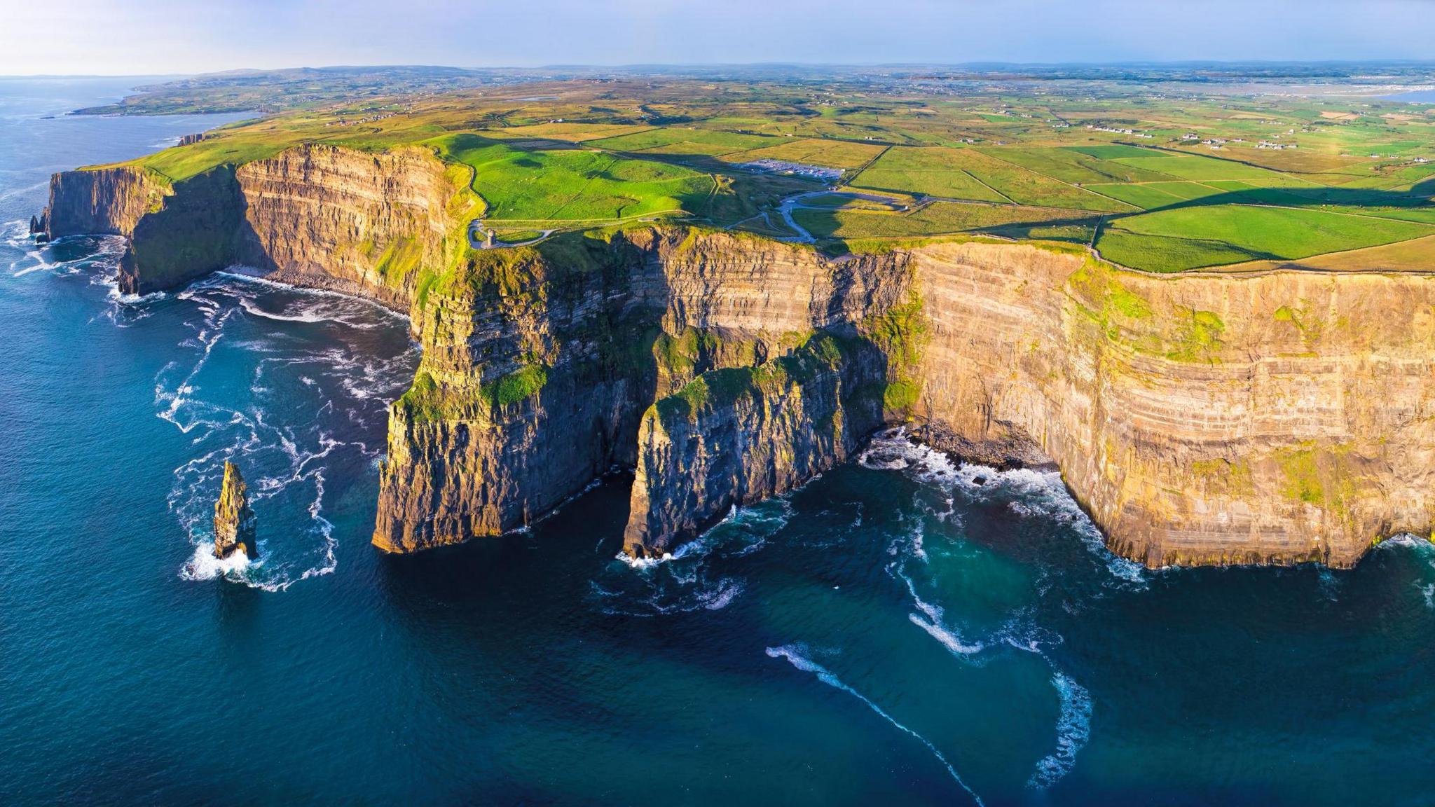 The Cliffs of Moher viewed from above on a sunny day. The photo is a panoramic view of the faces of the cliffs, with a calm sea below. Above the cliffs, flat green fields stretch into the distance.