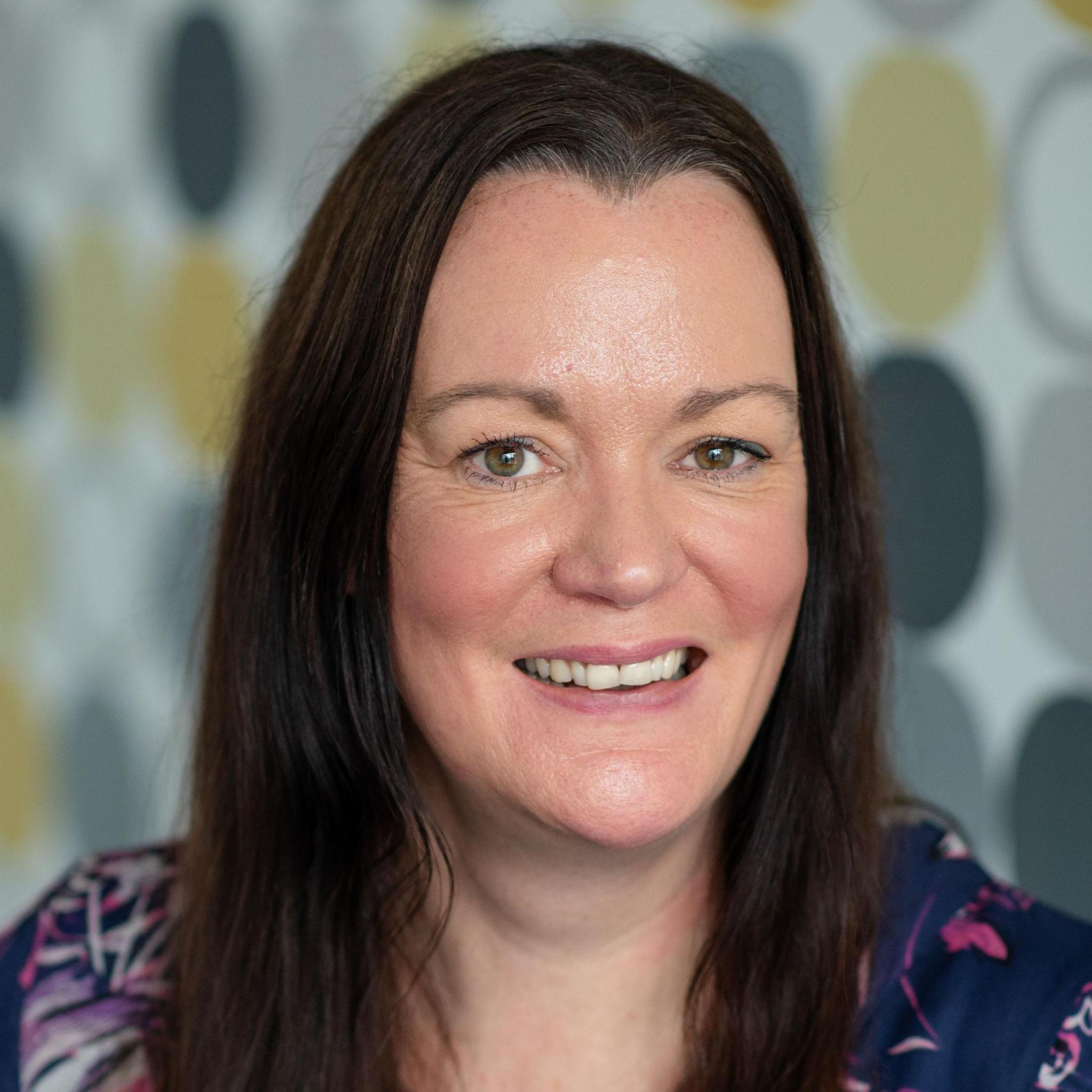 Kate Wallace, Victim Support Scotland. Woman with long dark hair, smiles looking into the camera wearing a navy blouse with pink and white details with a yellow and grey patterned wall out of focus behind her