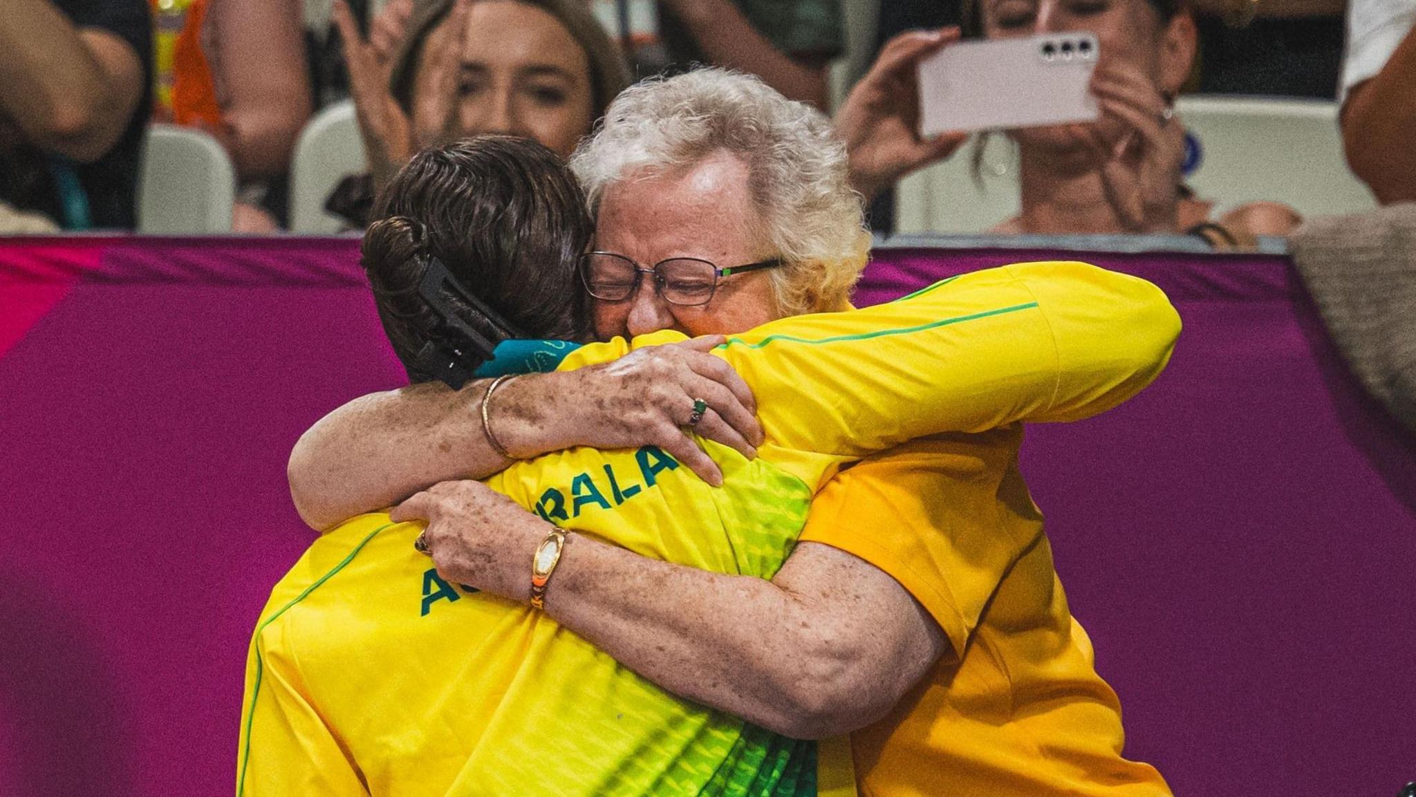 An athlete in a yellow and green tracksuit is hugged by a woman with grey hair, wearing an orange shirt.  The other spectators are warmly cheering and clapping.