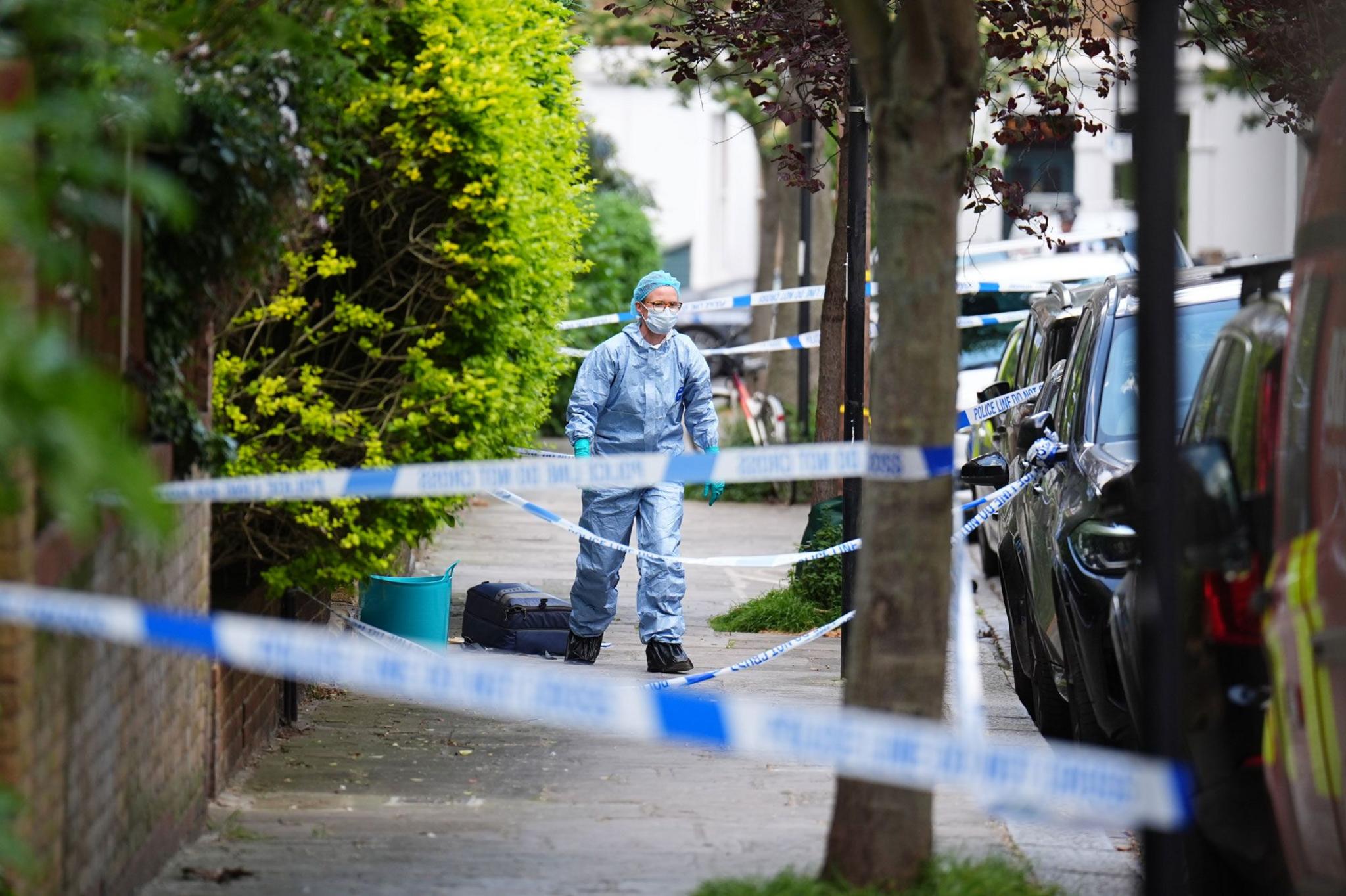 Police tape is tied across a street between trees as a forensic worker walks down it