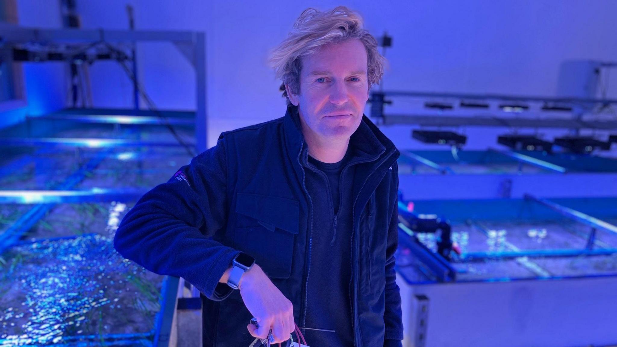 A man with blond hair standing in a blue lit room with tanks of water filled with seaweed in them.