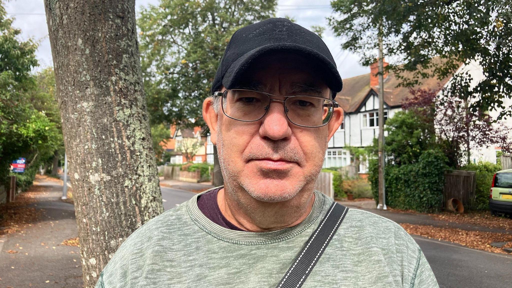 A head-and-shoulders photo of a man wearing a black baseball cap, glasses and a light green top looking straight at the camera. There's a tree-lined residential street behind him.