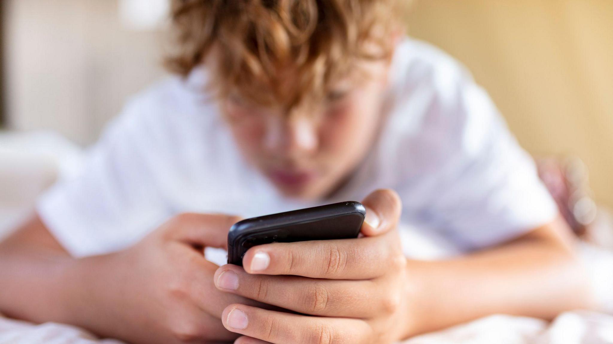 Fair-haired boy lying on his front looking at his mobile phone screen.