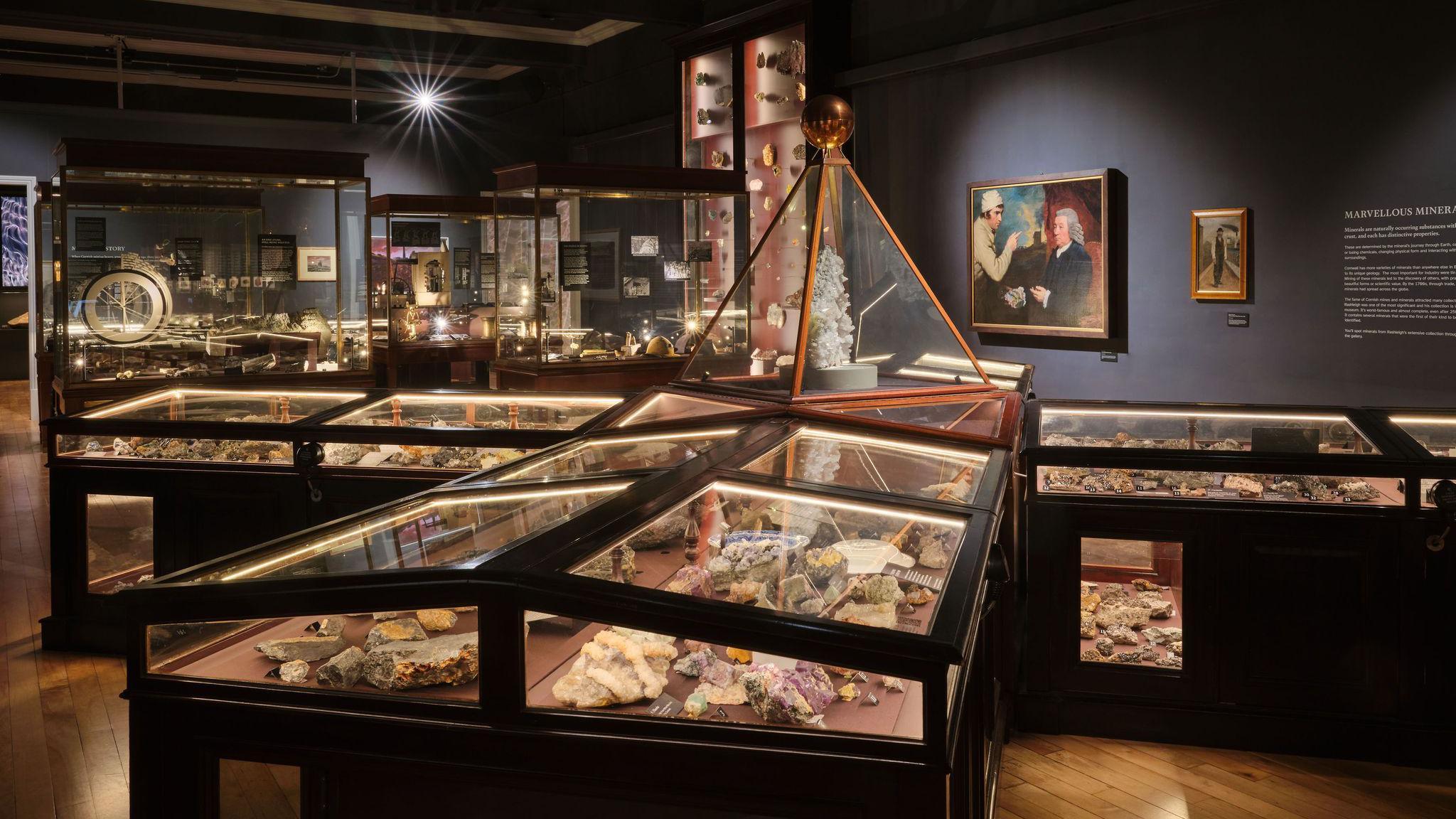 The photo shows an interior view of a museum exhibit. The room contains multiple glass display cases showcasing various rock specimens. The display cases are illuminated. There are more display cases in the background with some paintings on the wall. The room has wooden flooring and dark-coloured walls.