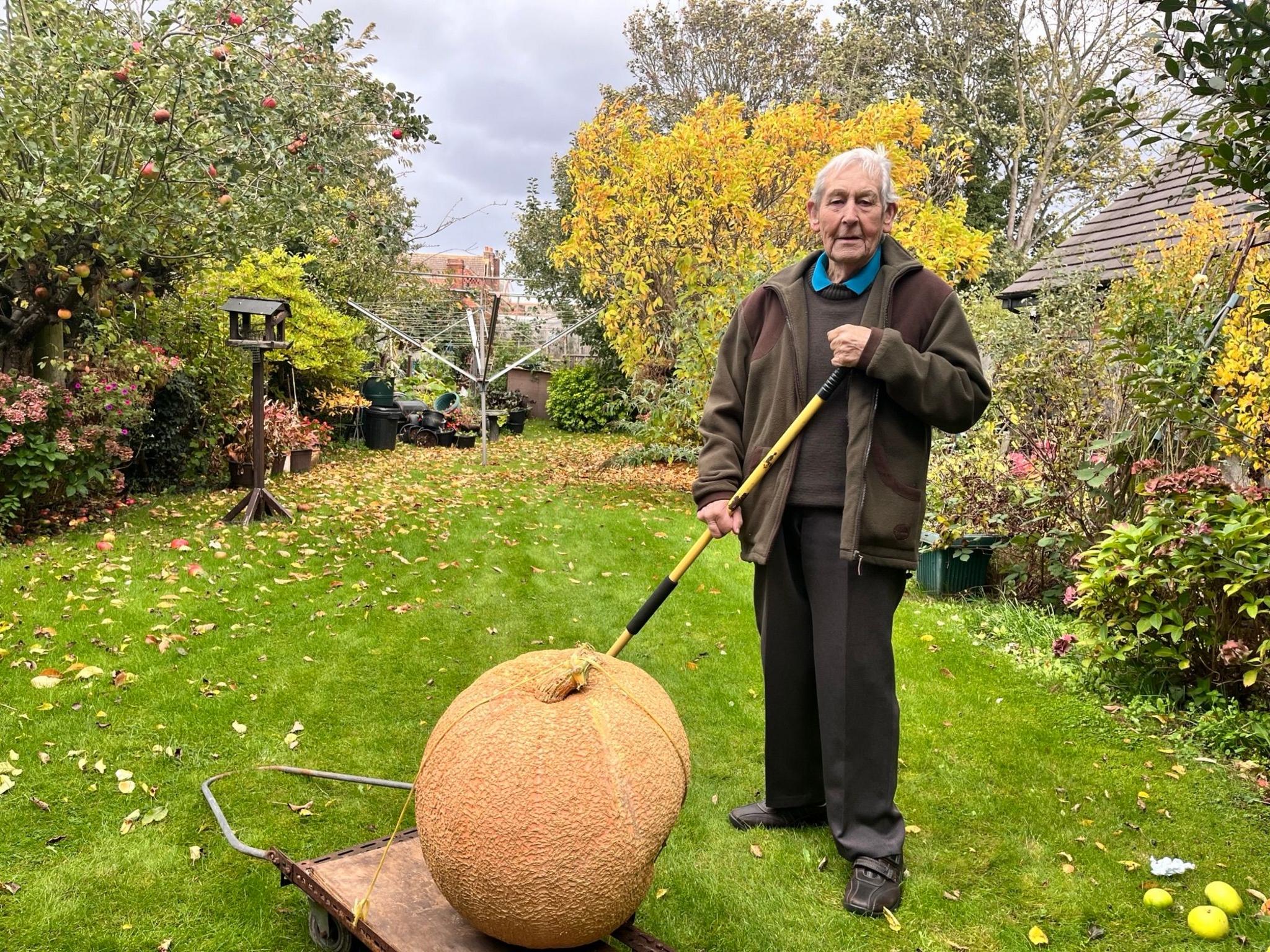 Fred Ramsey in his garden stood behind a large pumpkin. He has short grey hair and is wearing a green and brown coat, a black sweater, a blue collared t-shirt and dark-coloured trousers. Behind him is a bird house and a washing line.
