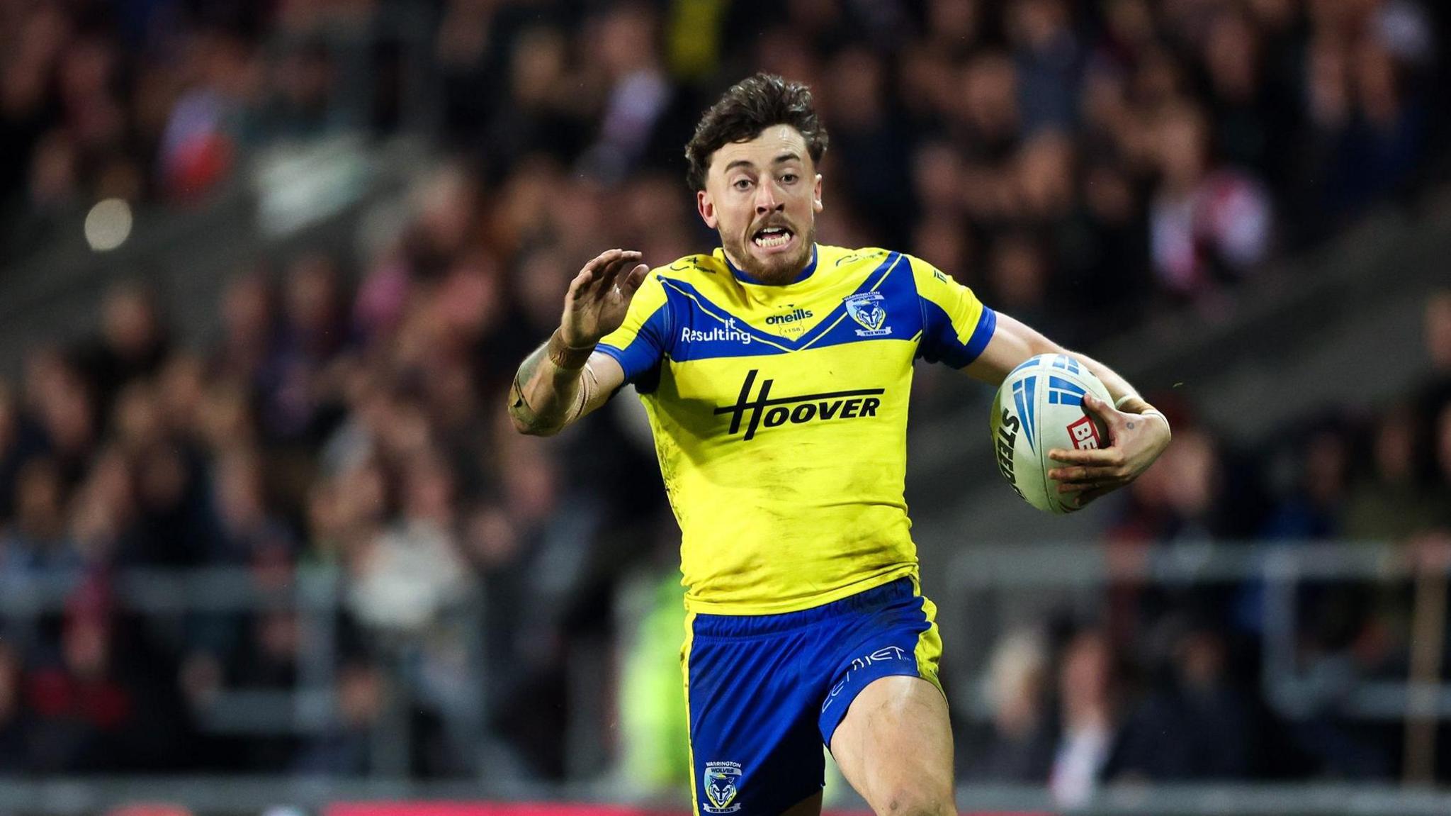 A photo of Matty Ashton running with the ball in his Warrington Wolves kit with the crowd in the background and an opposing player on the floor behind him