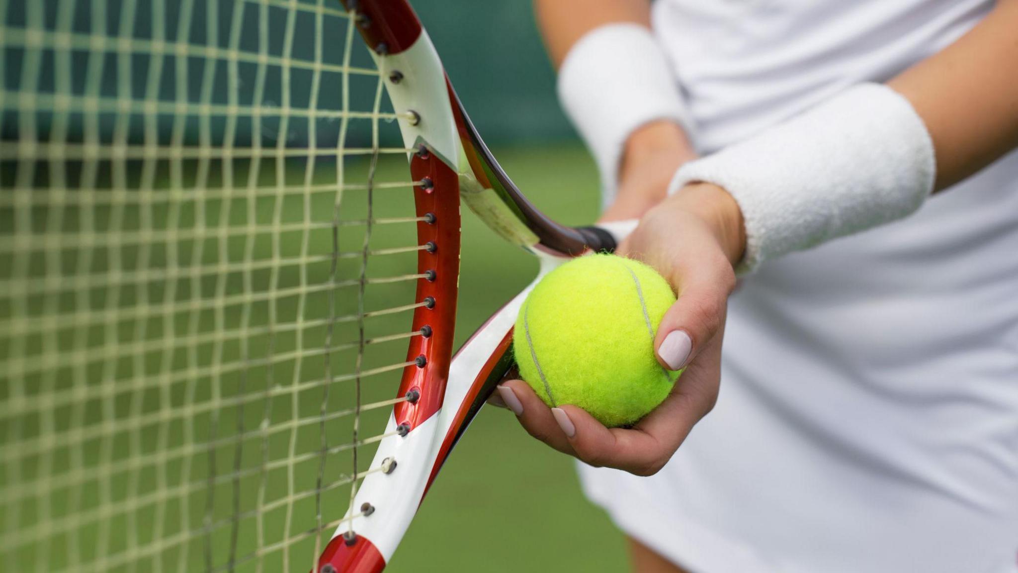 A woman in white holding a green tennis ball next to her red and white racket, getting ready to serve.