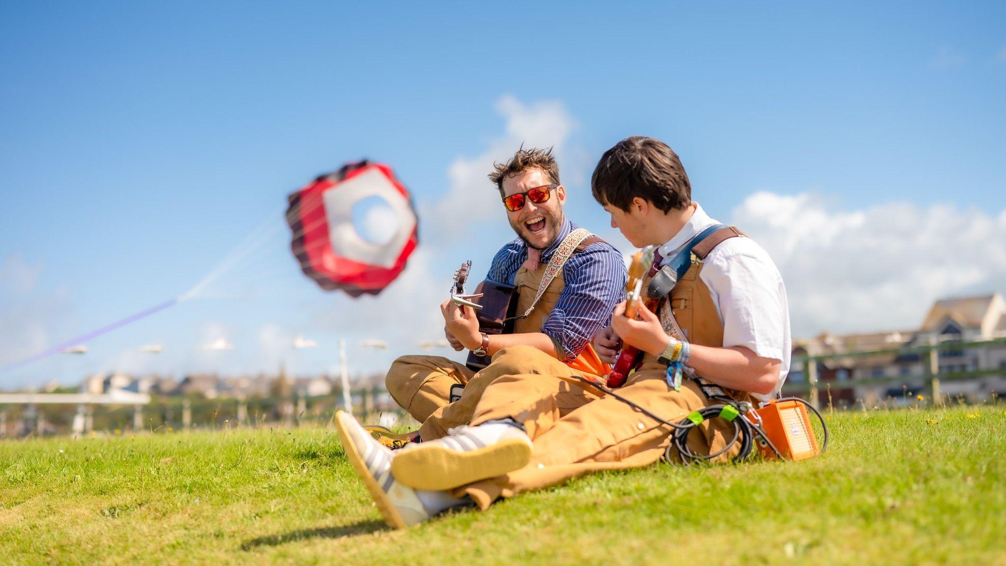 Two men play guitars as they sit on a grass verge in the sun. A large colourful kite is flying behind them.